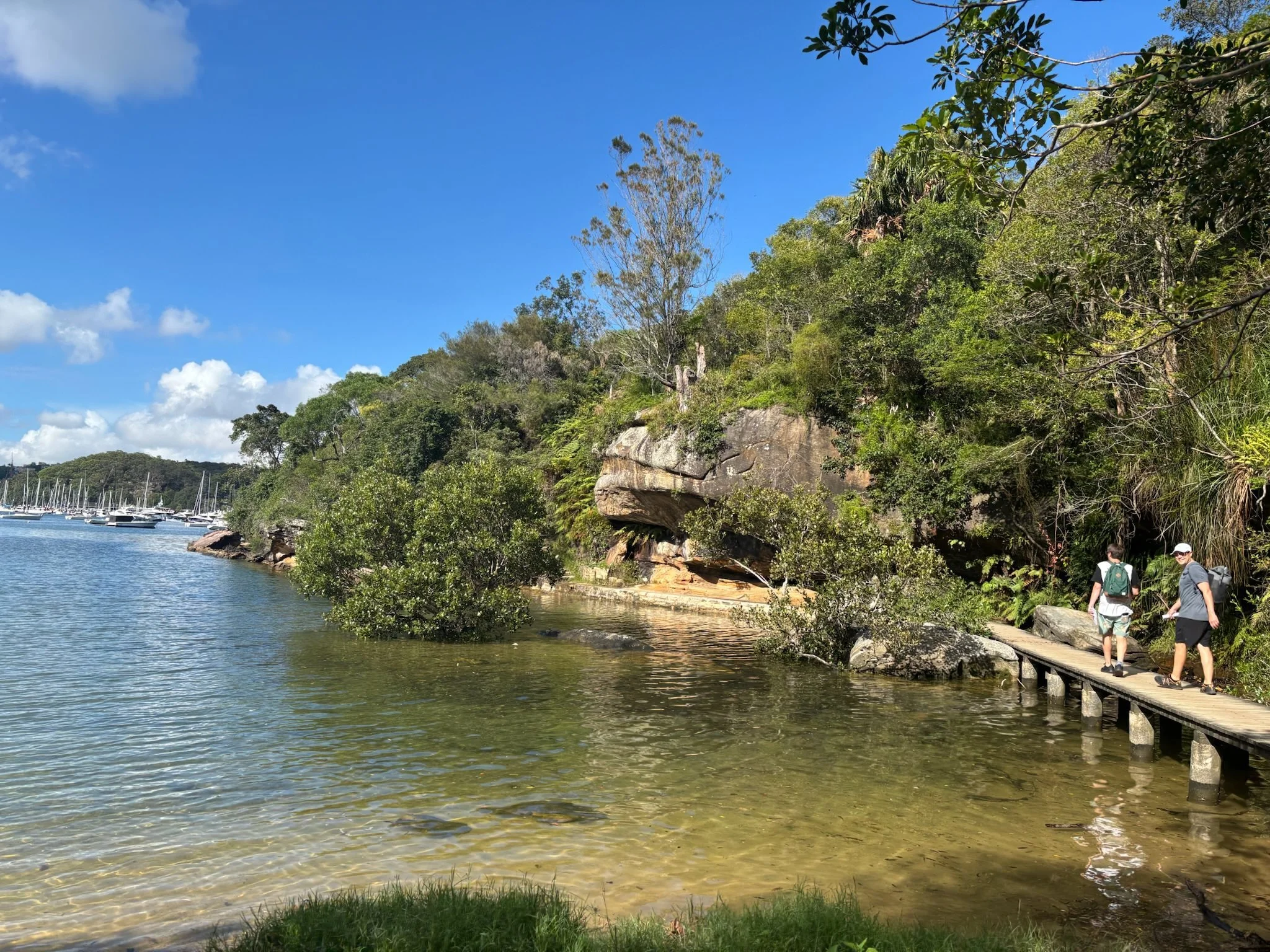 A tranquil Australian waterway with an overhanging rock formation and native trees provides a peaceful natural setting for leadership programs. Two people walk across a narrow bridge.