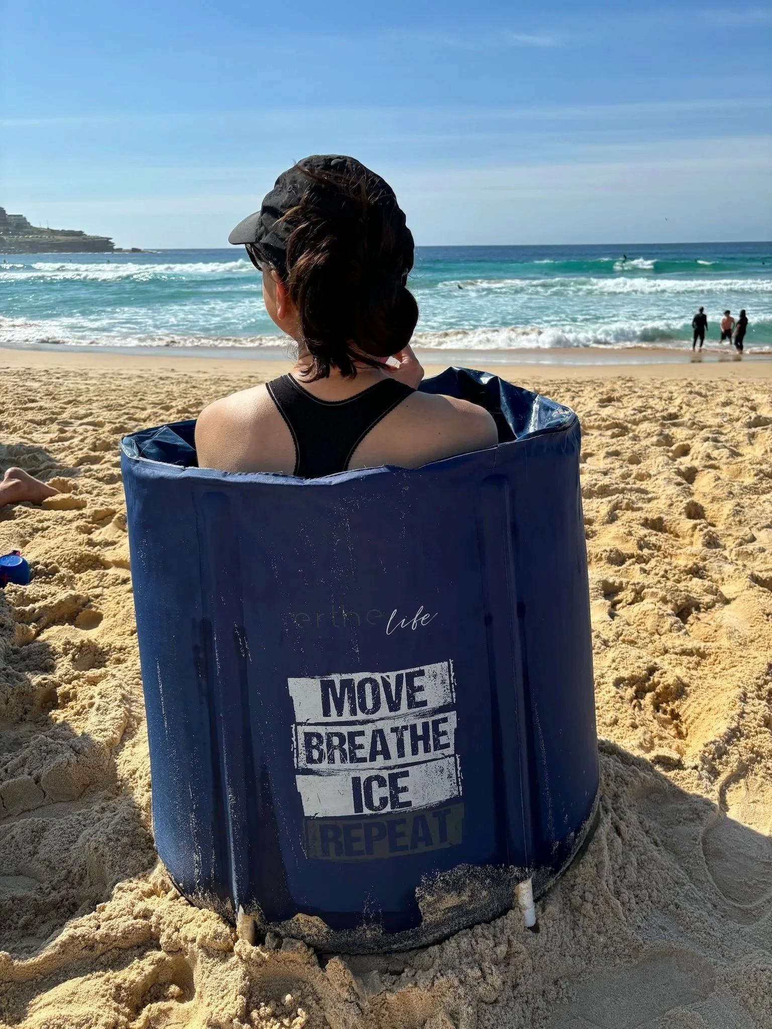 A leadership development program participant sits on a sandy beach facing the ocean. She is sitting in a blue cold plunge tub with 'MOVE BREATHE ICE REPEAT' text visibly printed on it.