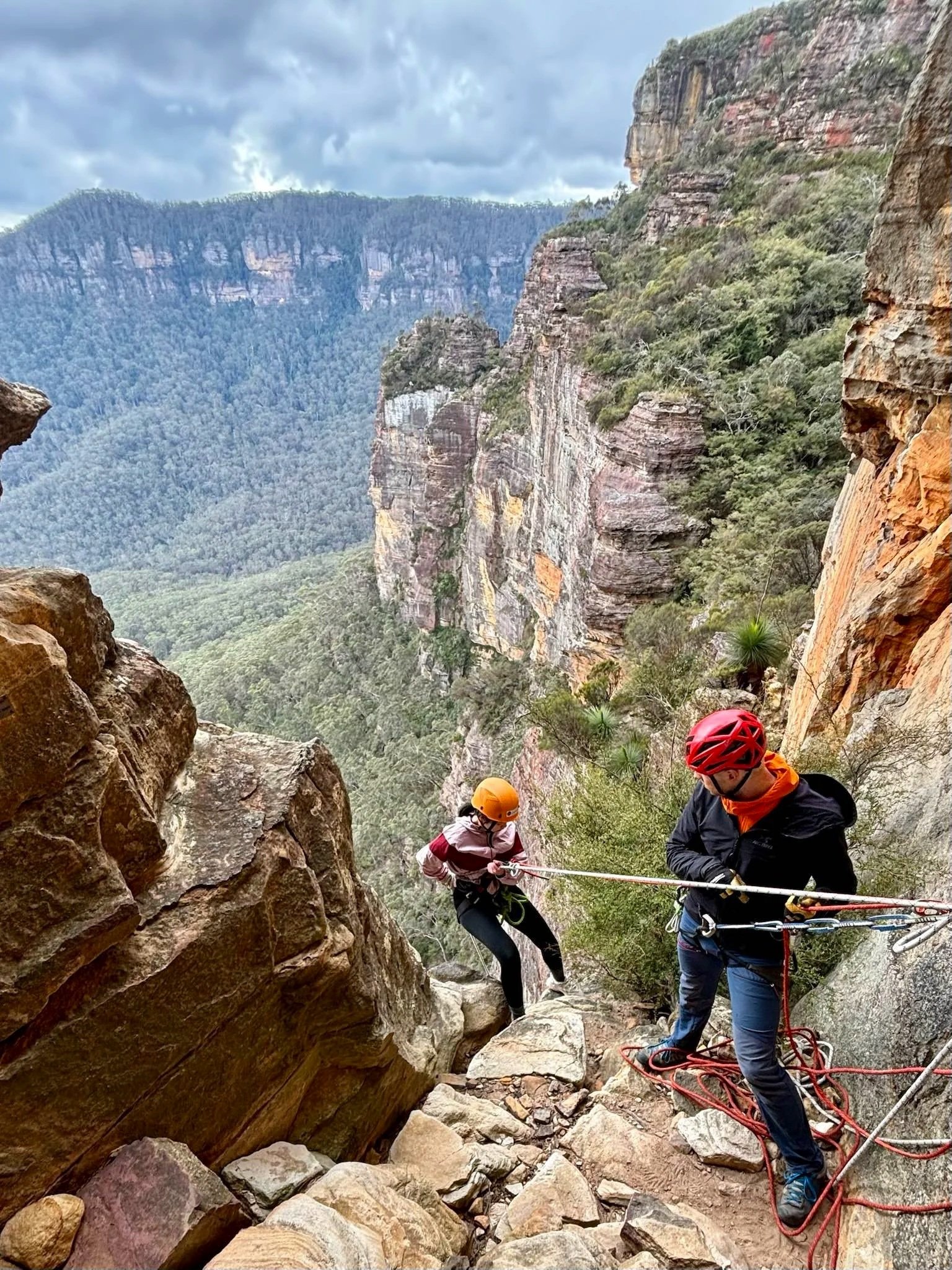 Participants navigate a rock climbing challenge during an outdoor leadership development program, using safety ropes on a cliff edge with dramatic canyon views in Australia.