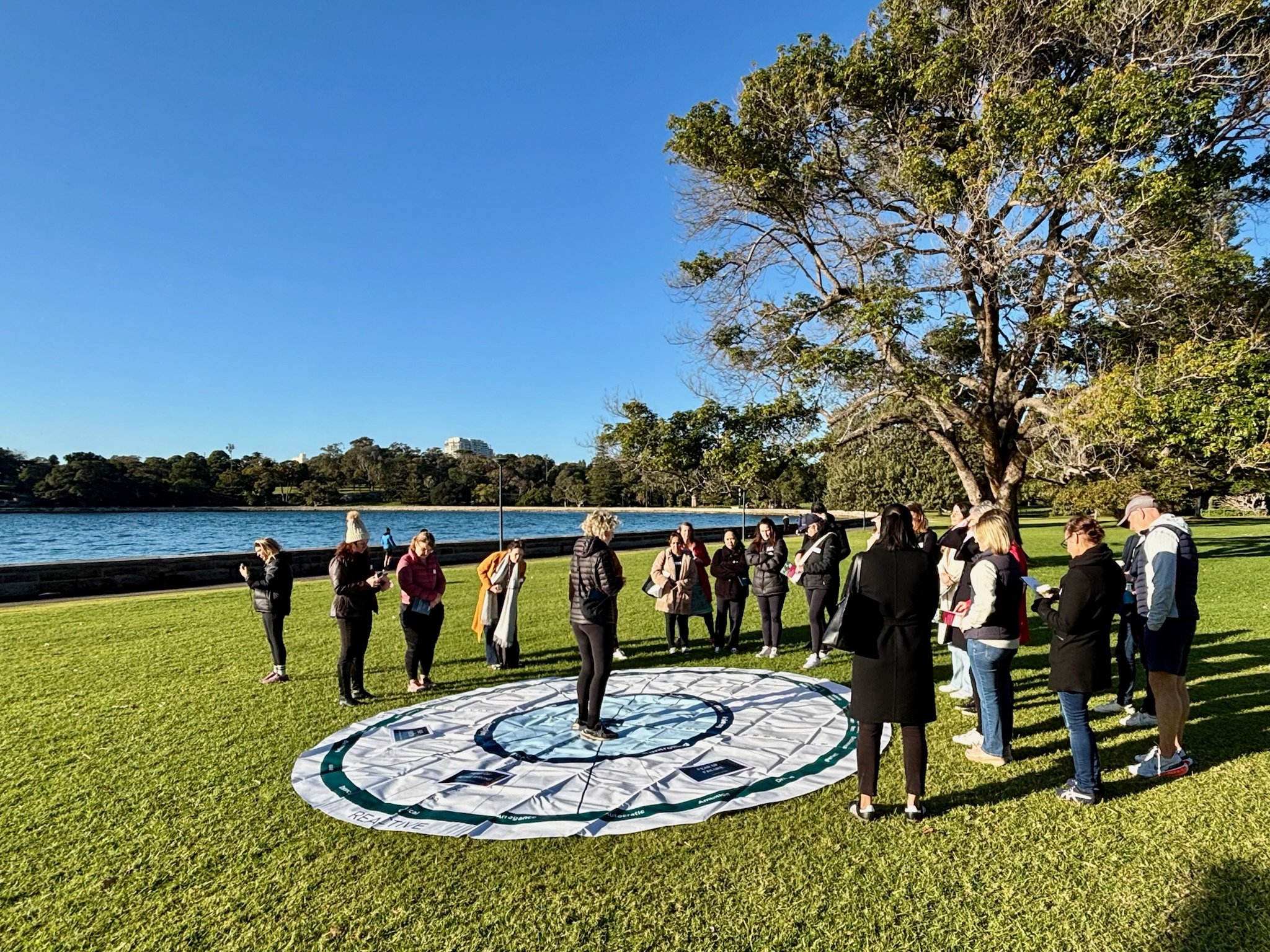 A team gathers around a large Leadership Circle diagram on a waterfront lawn during a Collective Leadership Lab, building trust and collaborative capability through shared experiences.