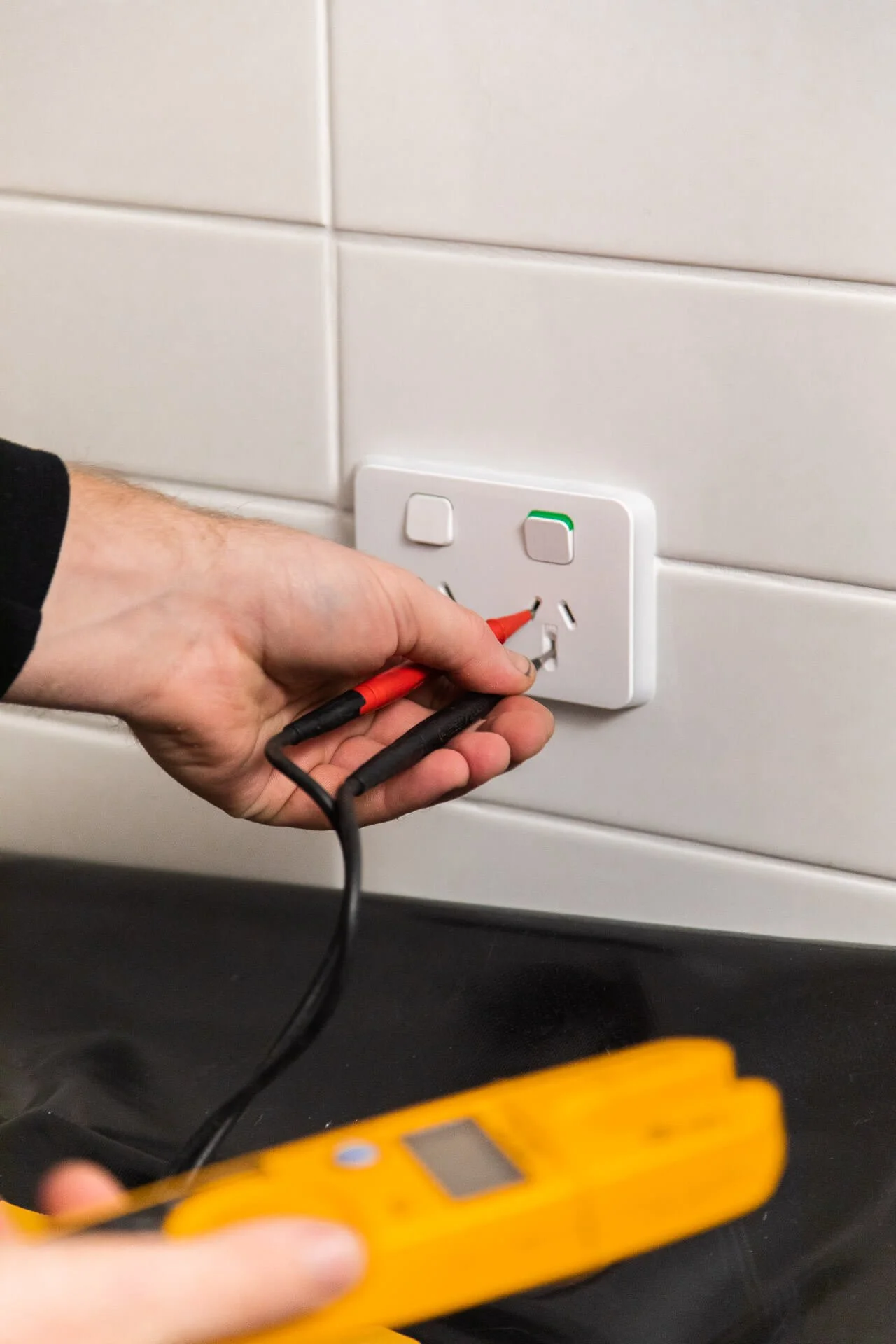 Person testing electrical socket with a multimeter in a room with white tile walls.