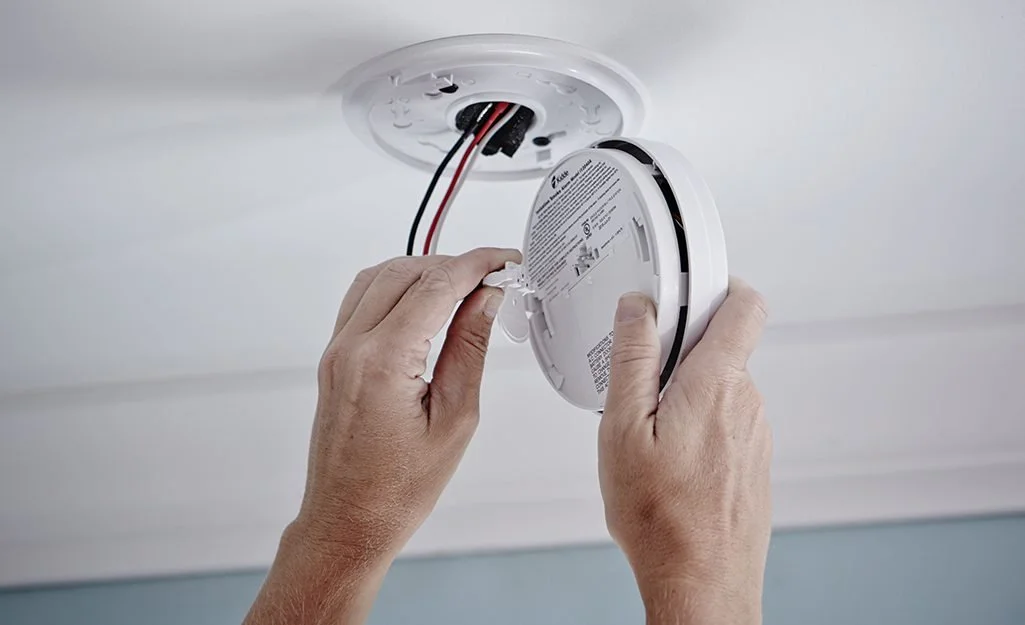 Person installing or replacing a smoke detector on the ceiling with wires exposed.