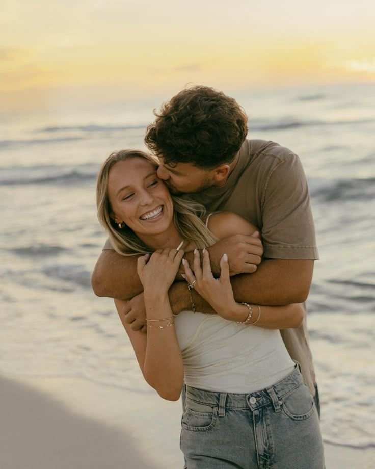 A couple hugging and sharing a kiss on the beach at sunset, with the woman smiling happily.