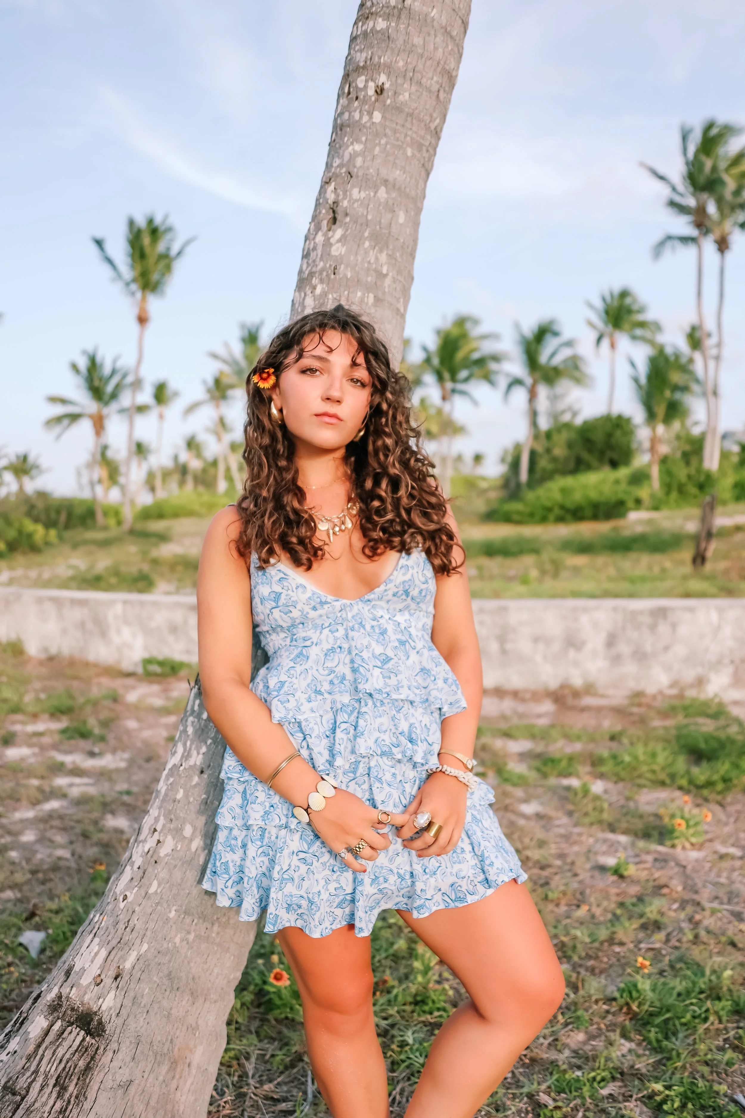 A young woman with curly hair and wearing a blue patterned dress, accessorized with jewelry, standing outdoors beside a leaning palm tree in a tropical setting with more palm trees in the background. In The  Bahamas on the island of Eleuthera.