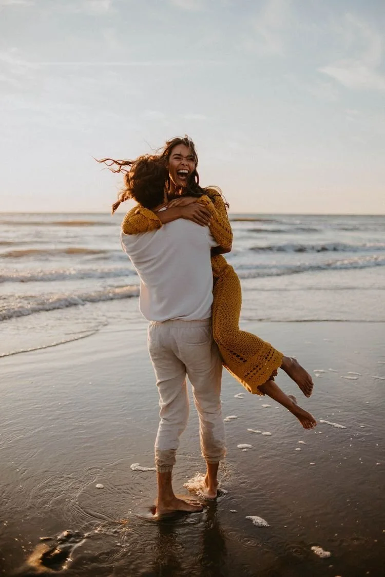 A person lifting a woman on the beach at sunset, both smiling and enjoying the moment.