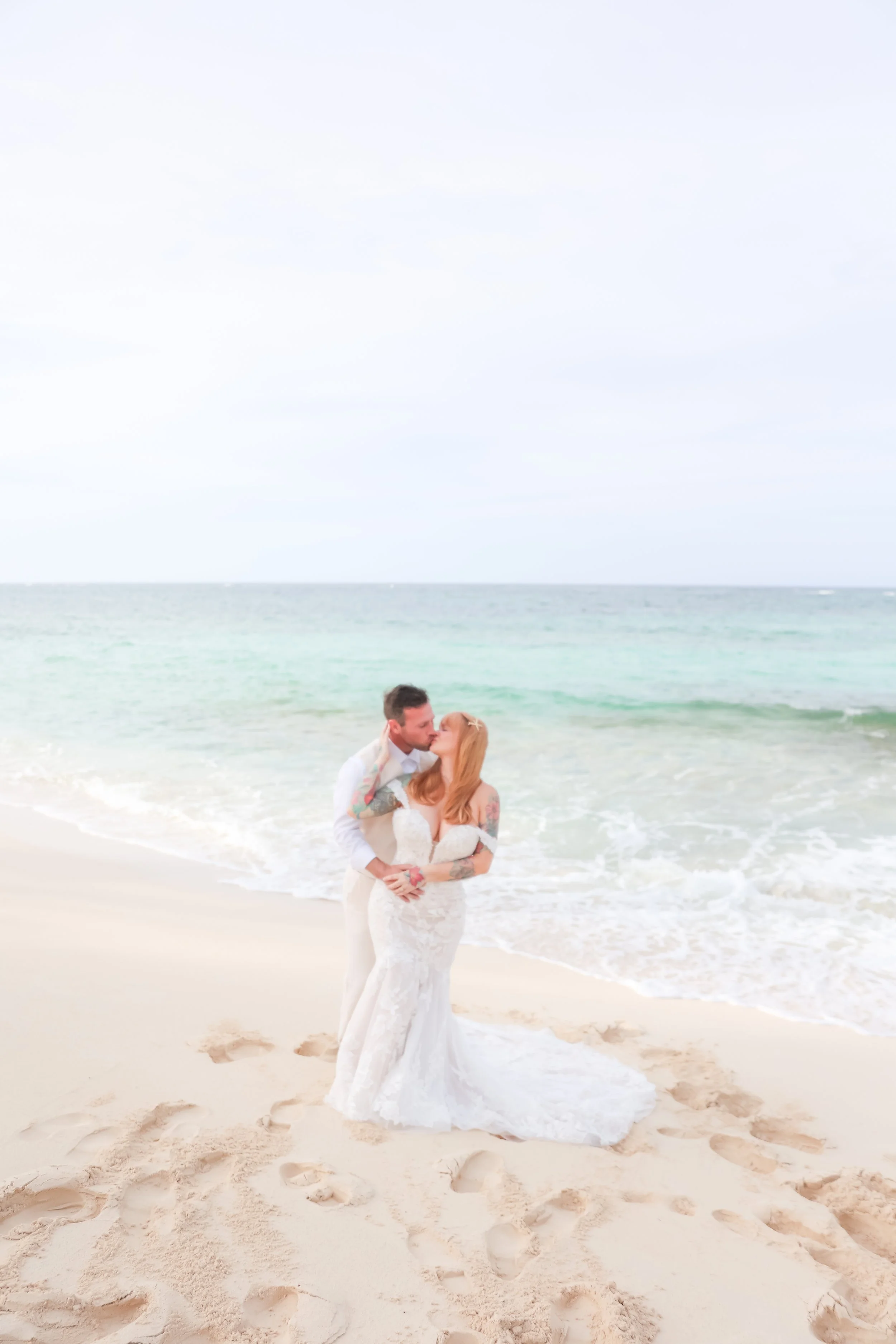 A couple in wedding attire, kissing on a sandy beach, with the ocean and cloudy sky in the background.