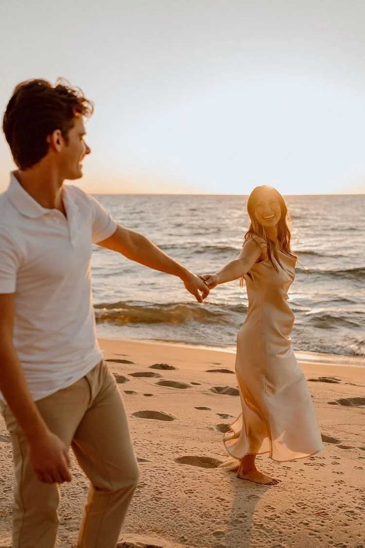 A couple holding hands on a beach during sunset, smiling and looking at each other.