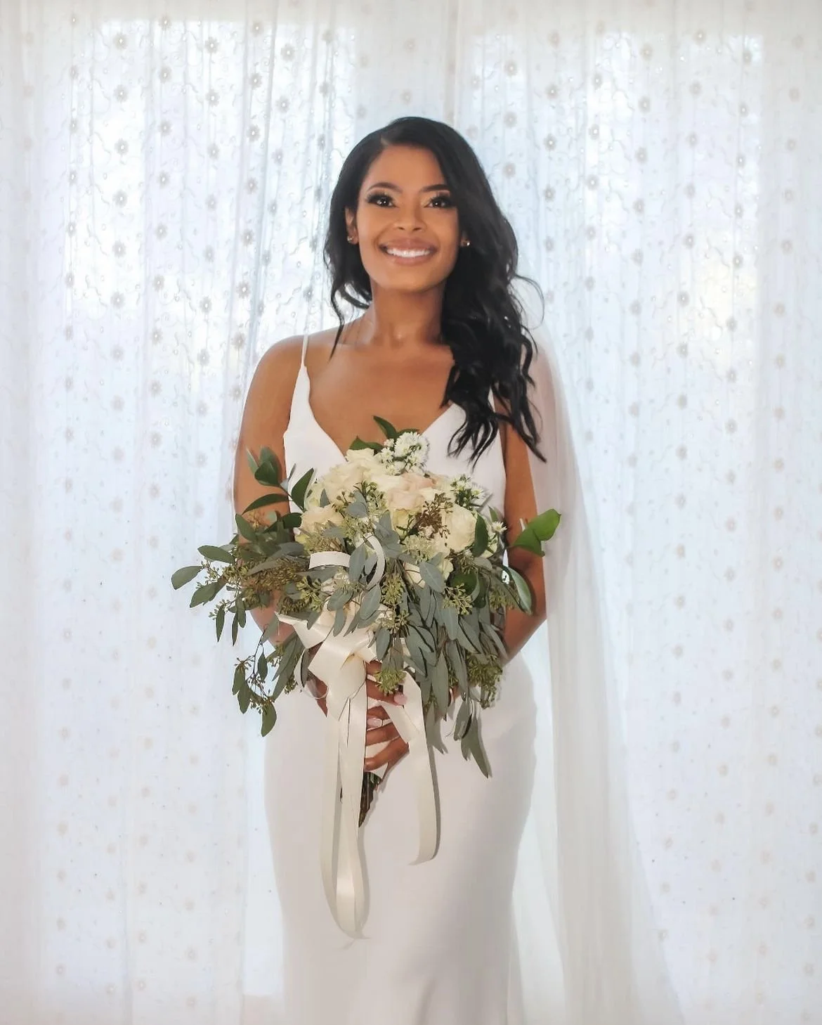 A woman with long black hair wearing a white dress, smiling, holding a bouquet of white and green flowers, in front of a light background with sheer curtains, wedding photography in Eleuthera