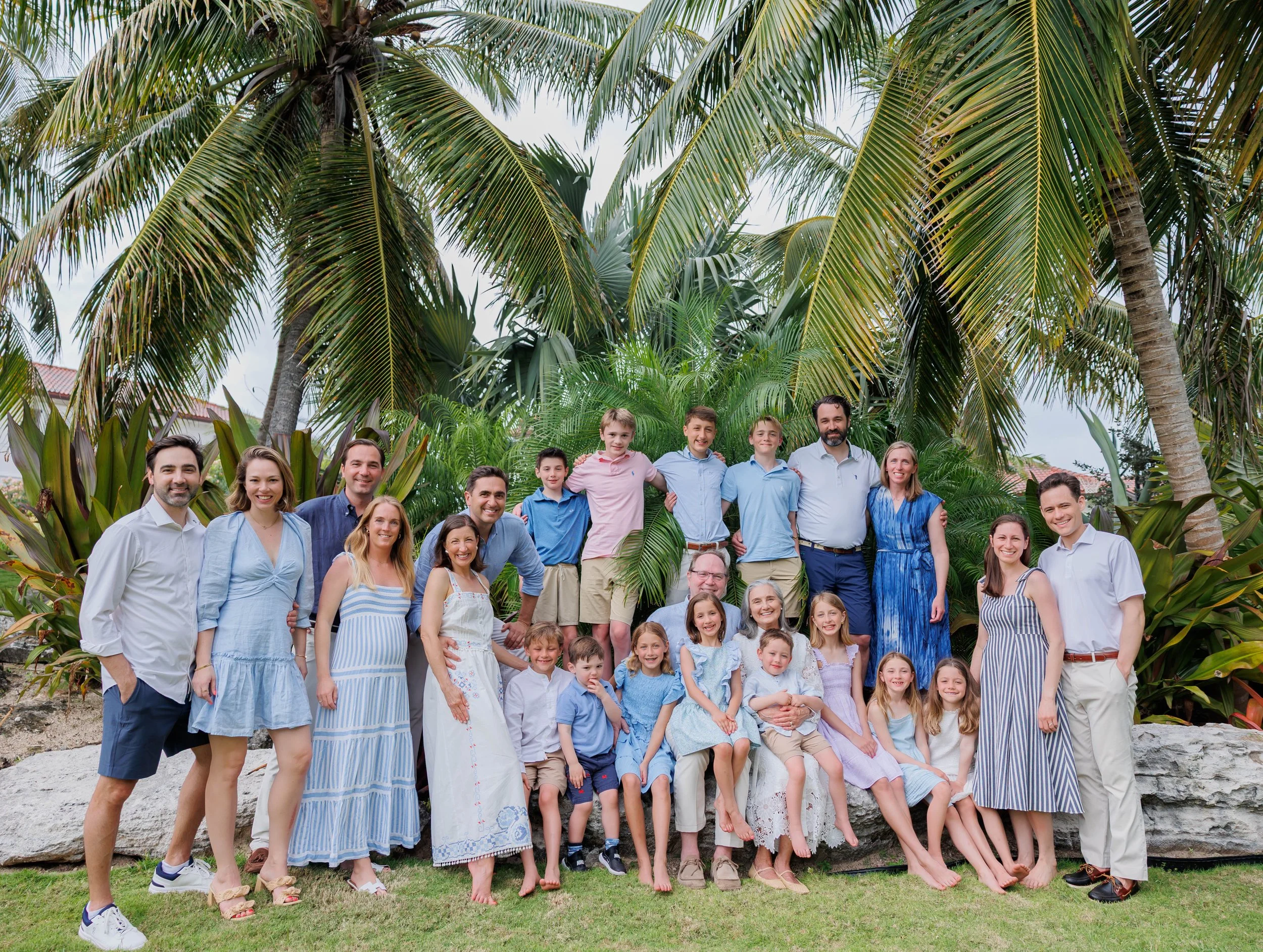 A large group of people, including adults and children, posing outdoors among tropical trees and plants, smiling for a photo.