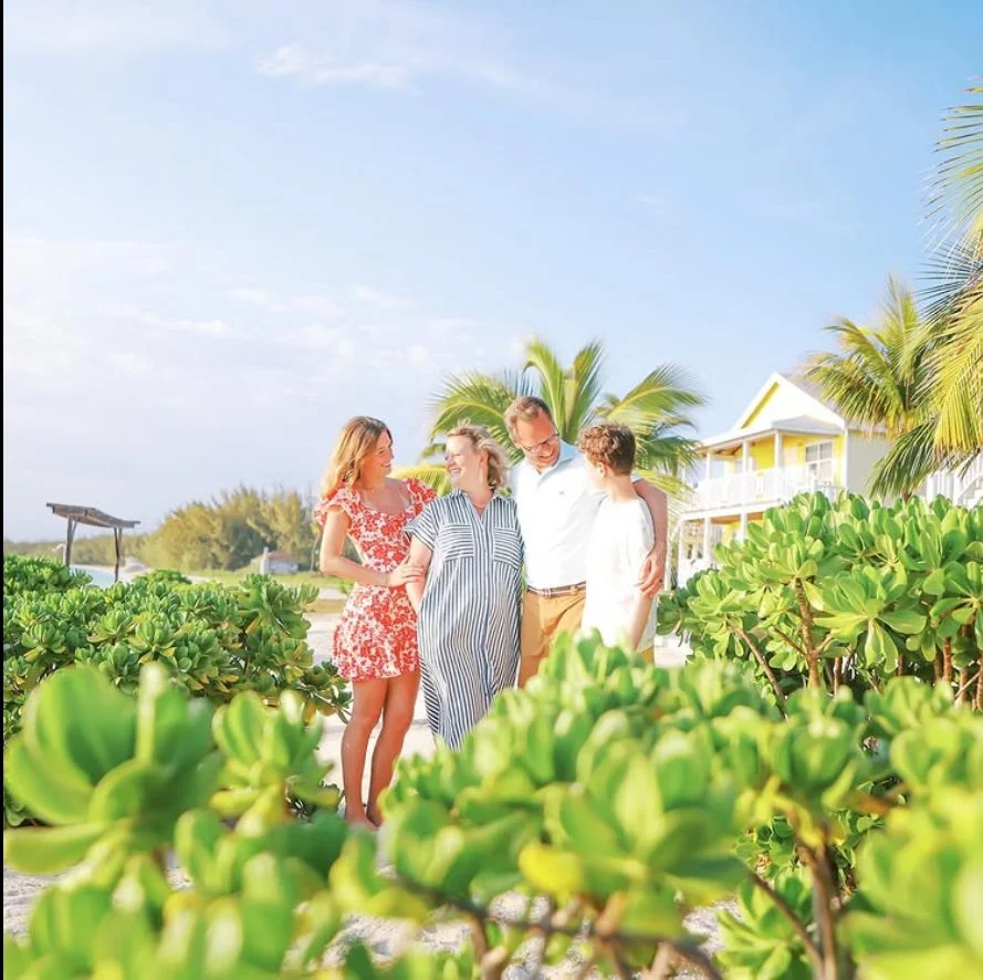 Family of four enjoying sunny day on beach with tropical plants, palm trees, and a white house with yellow accents in the background.