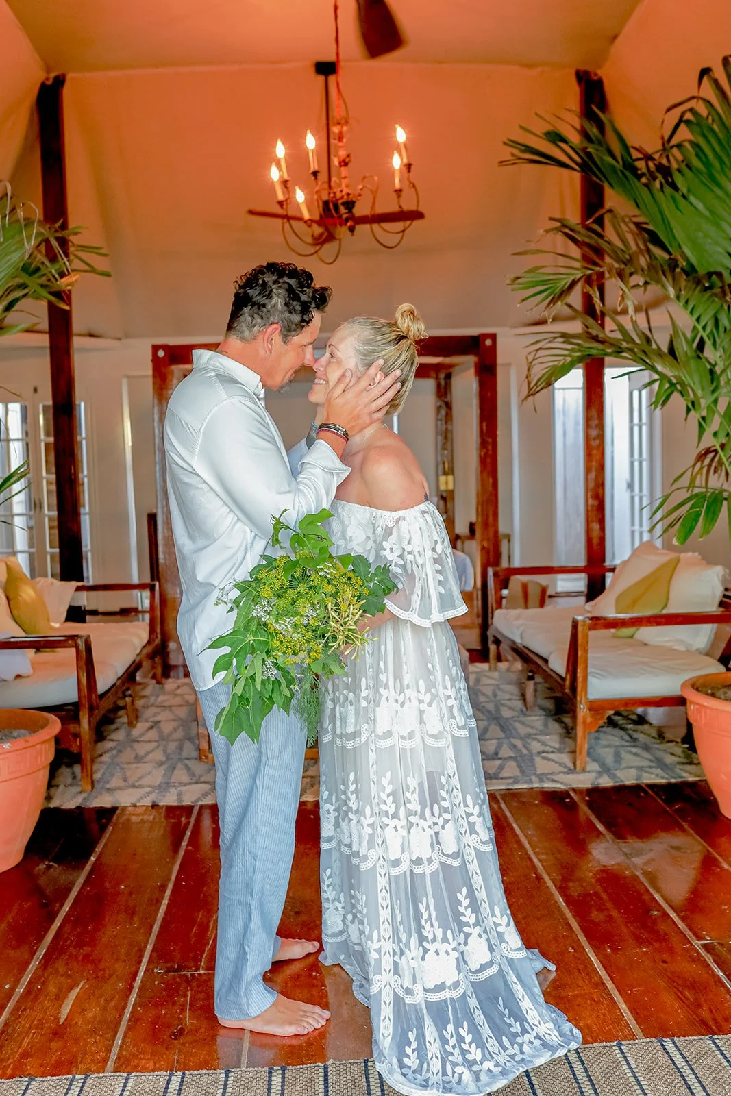 A couple in love, standing close and touching foreheads, in a cozy indoor setting with wooden furniture, potted plants, and a chandelier. bahamas wedding