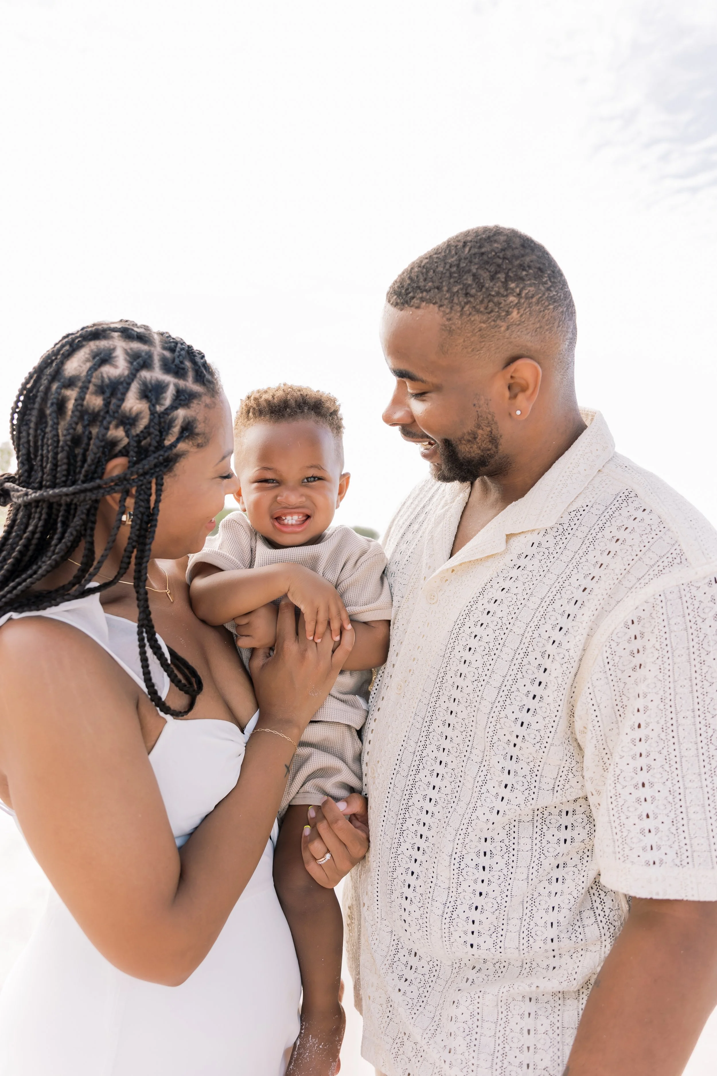 A happy family of three smiling on a sunny day, with a woman holding a young child and a man standing close.
