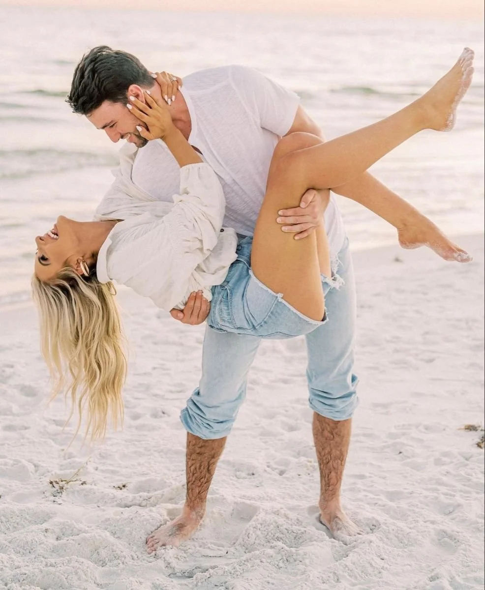 A man and woman are on a beach, with the man lifting and holding the woman upside down. They are smiling and appear to be enjoying each other's company.