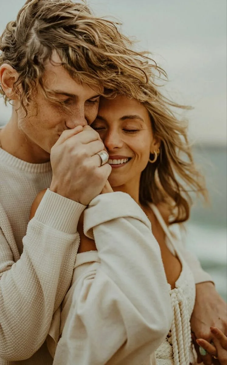A young man and woman sharing a tender moment, with the man gently kissing the woman's hand and both smiling contentedly.