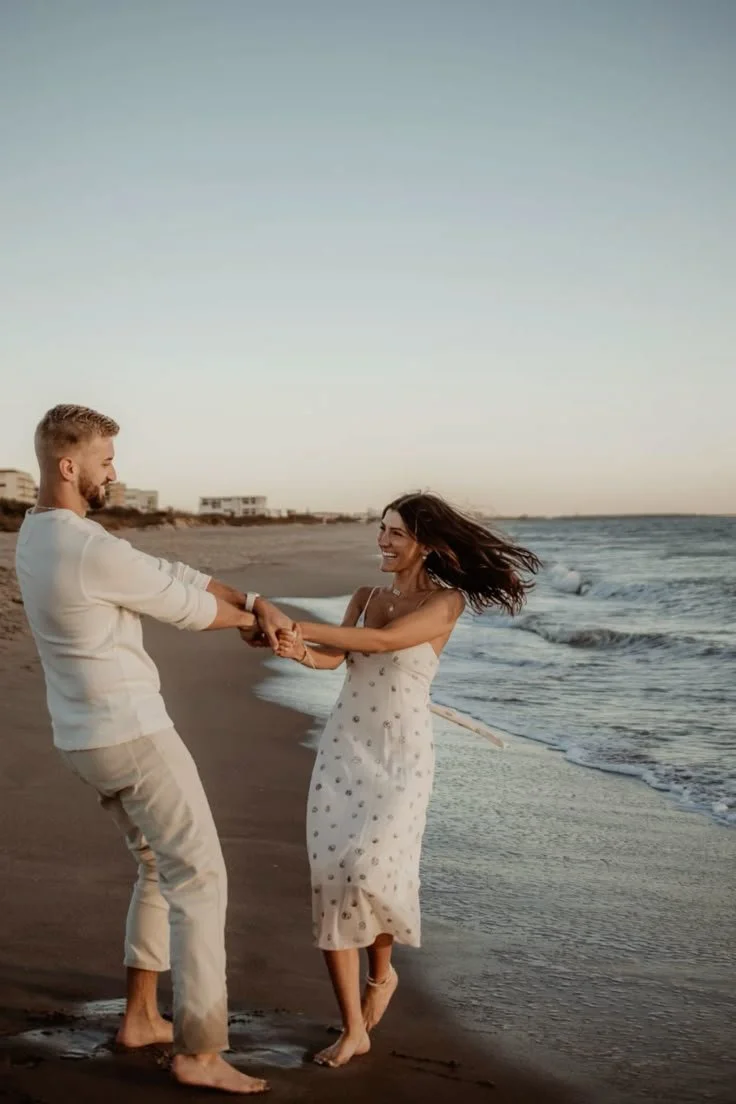 A couple holding hands and dancing on the beach with ocean waves in the background and buildings in the distance.