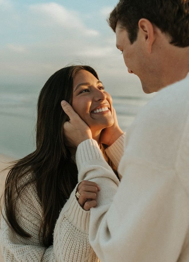 A smiling woman with long dark hair looks up at a man, who is holding her face and looking at her affectionately, while standing on a beach with the ocean in the background.