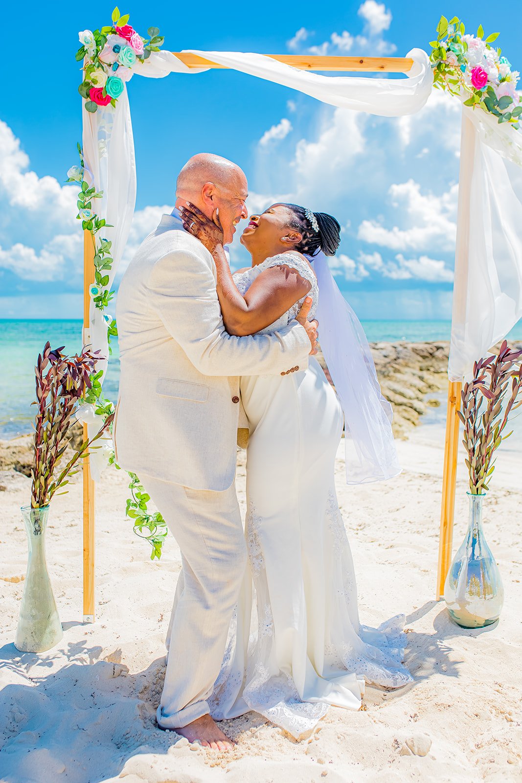 A couple in wedding attire celebrating their wedding on a beach, standing under a decorated wooden arch with flowers, with the ocean and sky in the background.