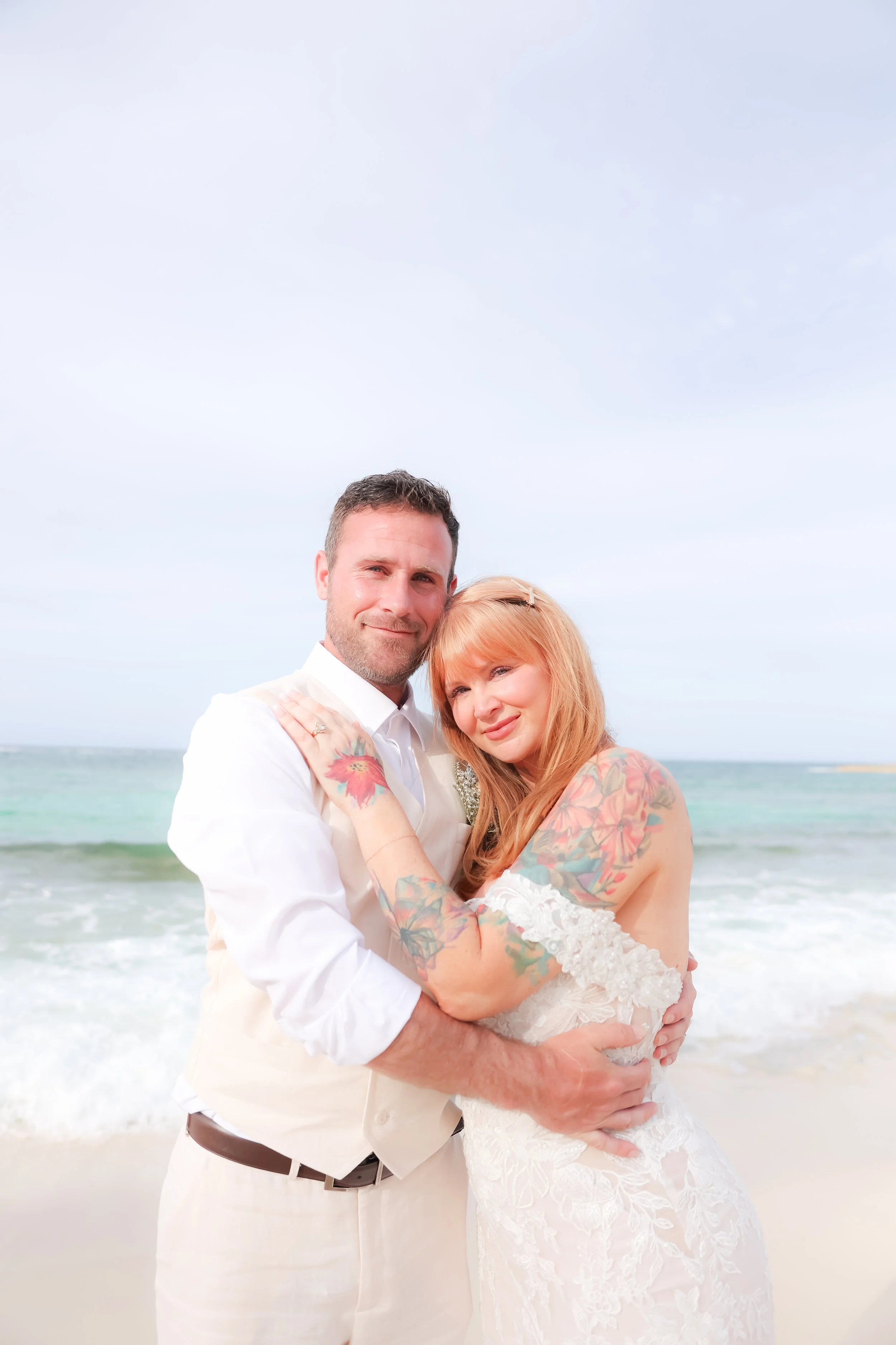 A couple in white wedding attire stands on a beach, embracing each other with in front of the ocean and a cloudy sky, wedding photography in Eleuthera