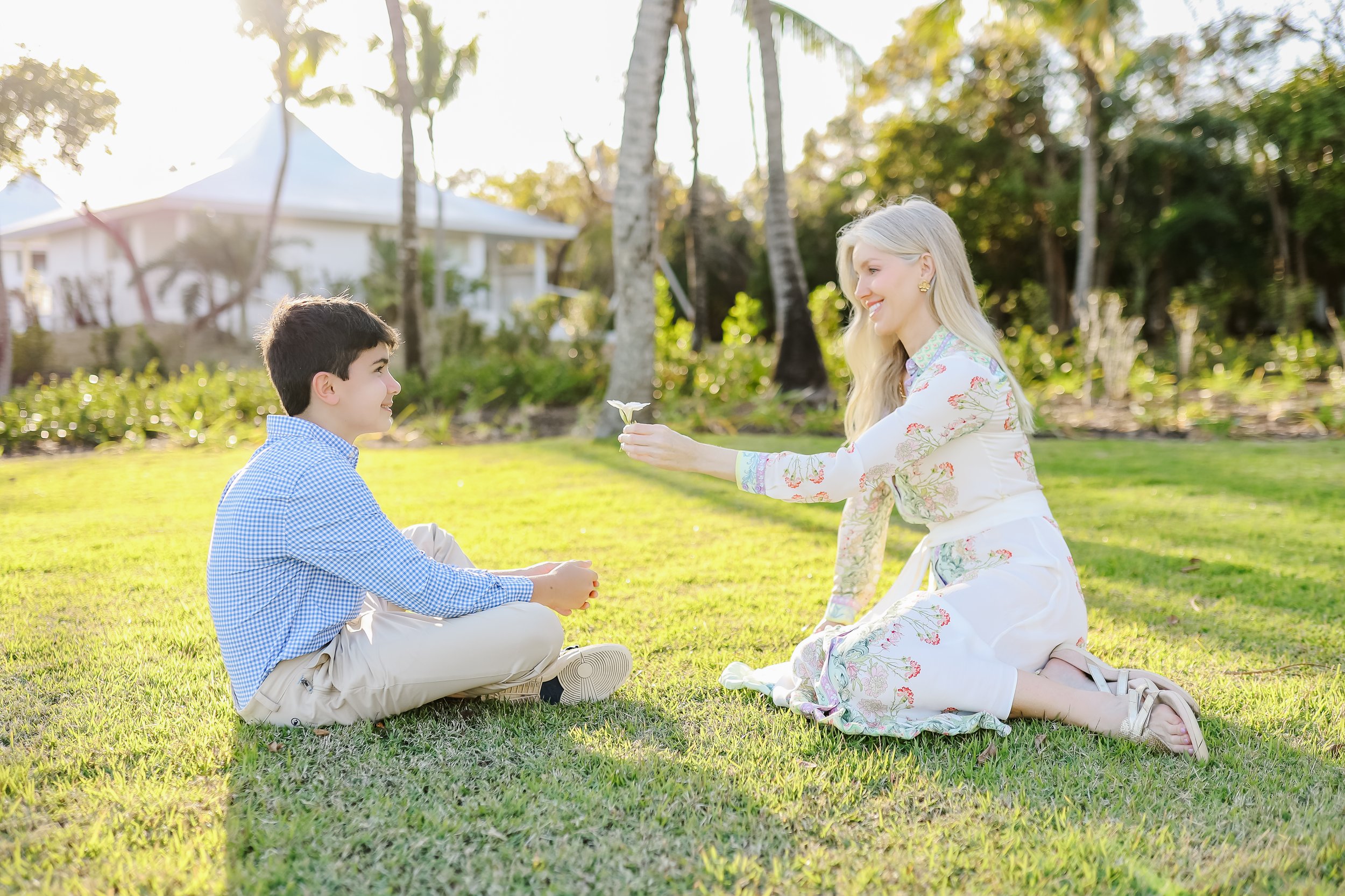 A woman and a young boy outdoors on a sunny day, i  with the woman kneeling on the grass and offering a flower to the boy, who is sitting cross-legged. They are surrounded by trees and a house in the background in.Family  photography in Eleuthera