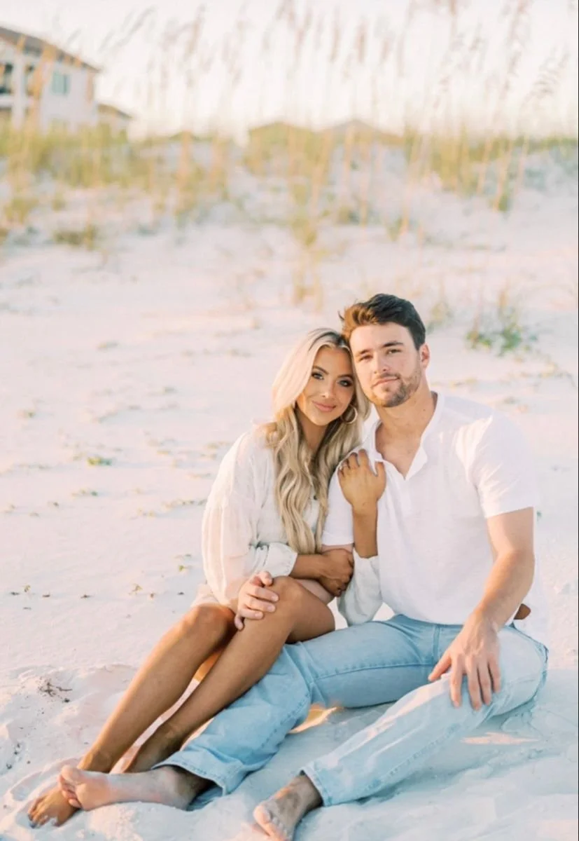 A couple sitting together on the sandy beach with the woman leaning on the man's shoulder, both smiling at the camera, in a bright outdoor setting.
