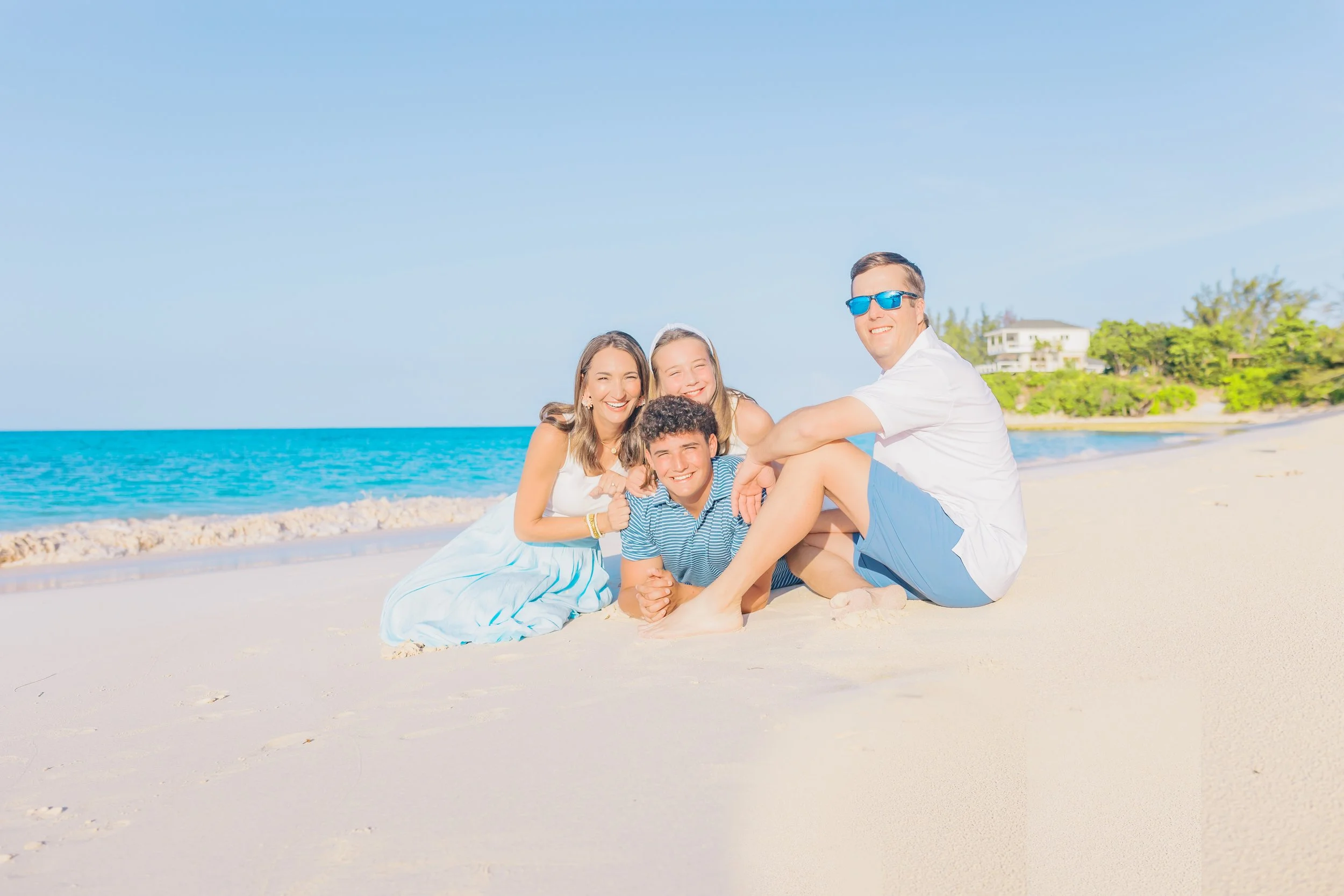 A family of four enjoying a sunny day on the beach, sitting on the sand with the ocean and a house surrounded by greenery in the background.
