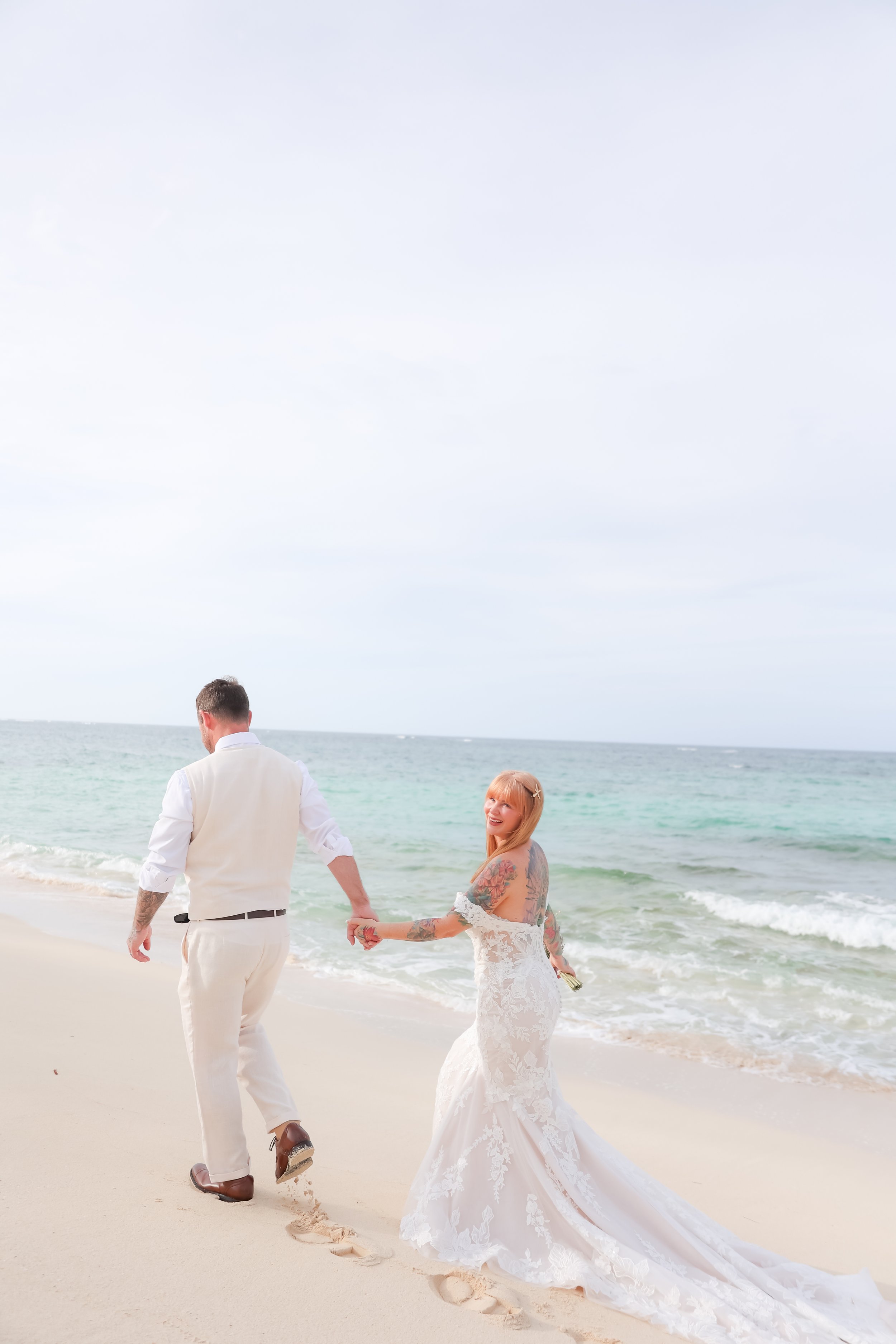 A newlywed couple holding hands and walking on a sandy beach near the ocean, with the bride wearing a white lace wedding dress and the groom in a light-colored suit,wedding photography in Eleuthera