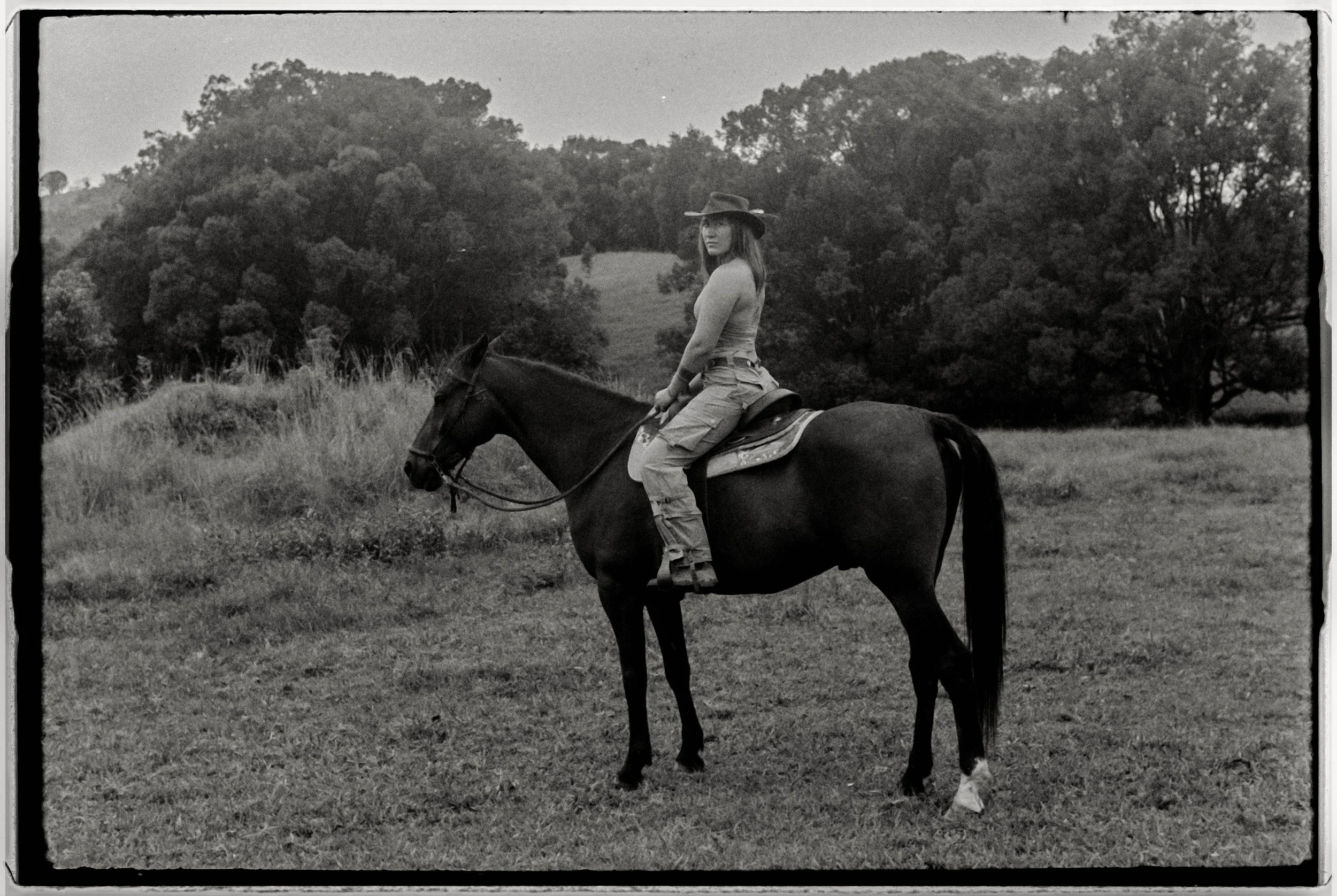 A woman wearing a hat and casual clothes riding a horse across a grassy field with trees in the background.