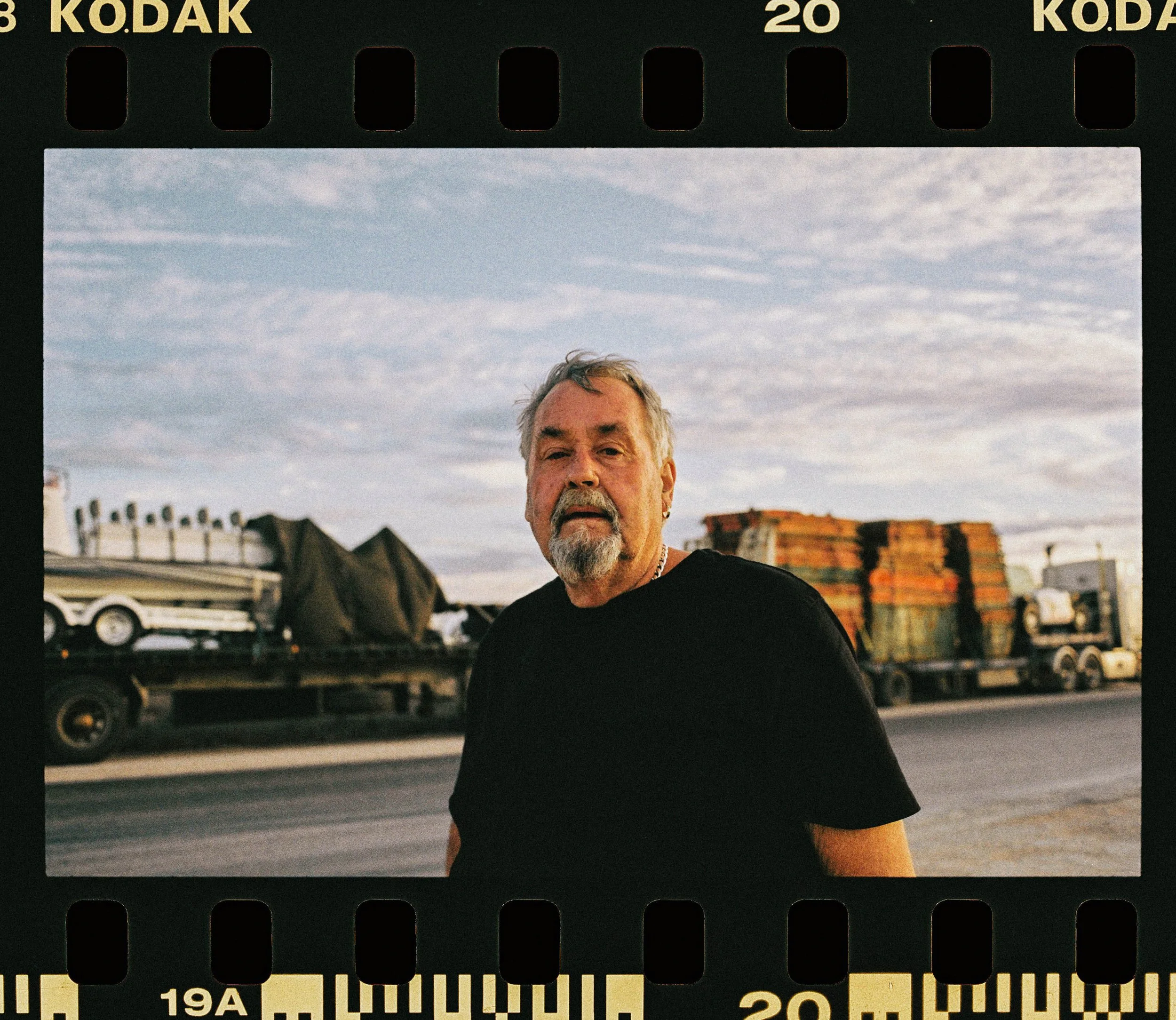 The image shows an older man with gray hair and a beard, standing outdoors against a background of a blue sky with some clouds and a cityscape with buildings and parked cars. The photo is bordered with black film strip edges with the word 'KODAK' printed at the top.