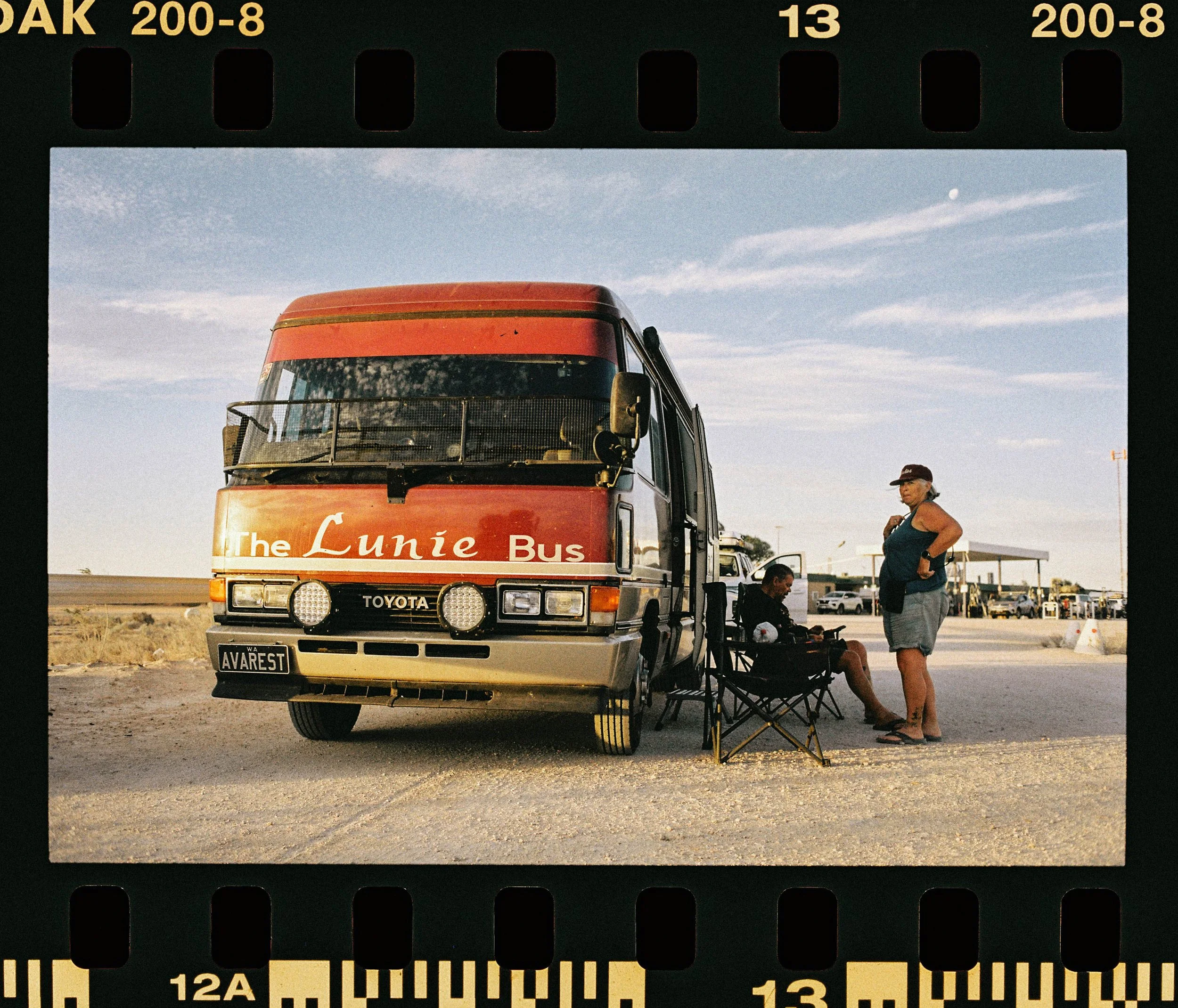 A red bus labeled 'The Lunnie Bus' parked outdoors with people nearby, including a woman in a blue top and hat, and two individuals sitting on a foldable chair. The scene is under a sky with scattered clouds and a visible moon.