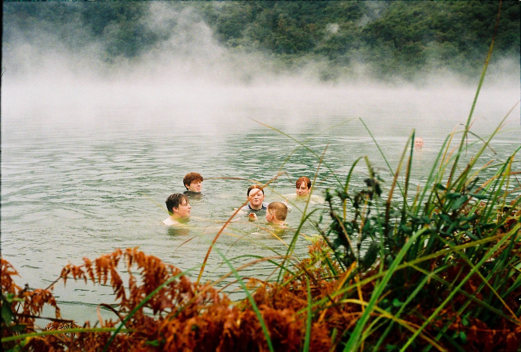 Group of people swimming and wading in a foggy lake surrounded by greenery and plants.