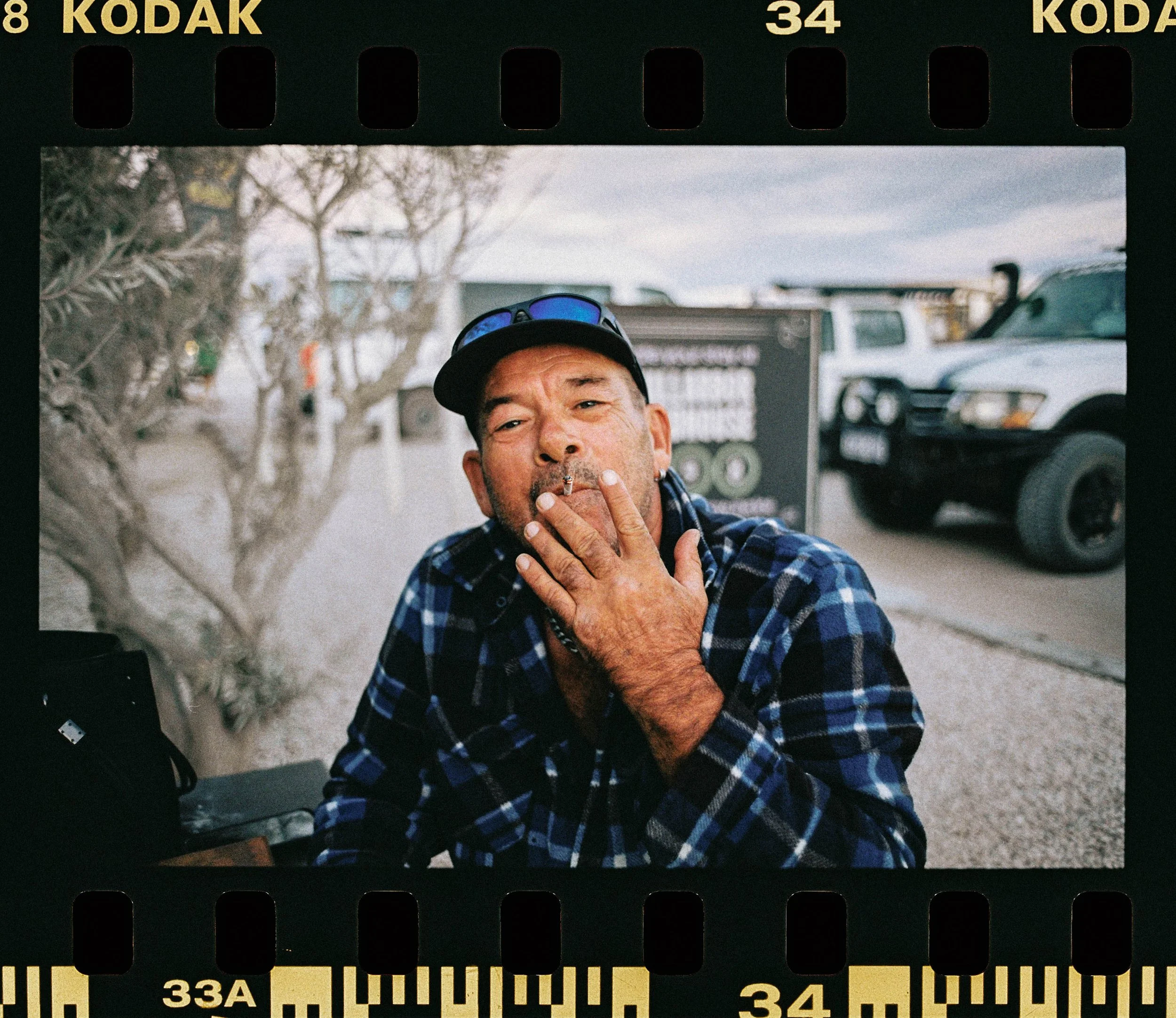 Close-up view of a man with a cigarette in his hand, wearing a blue checkered shirt, sunglasses on his head, and a black cap, standing outdoors with cars and blurry trees in the background.