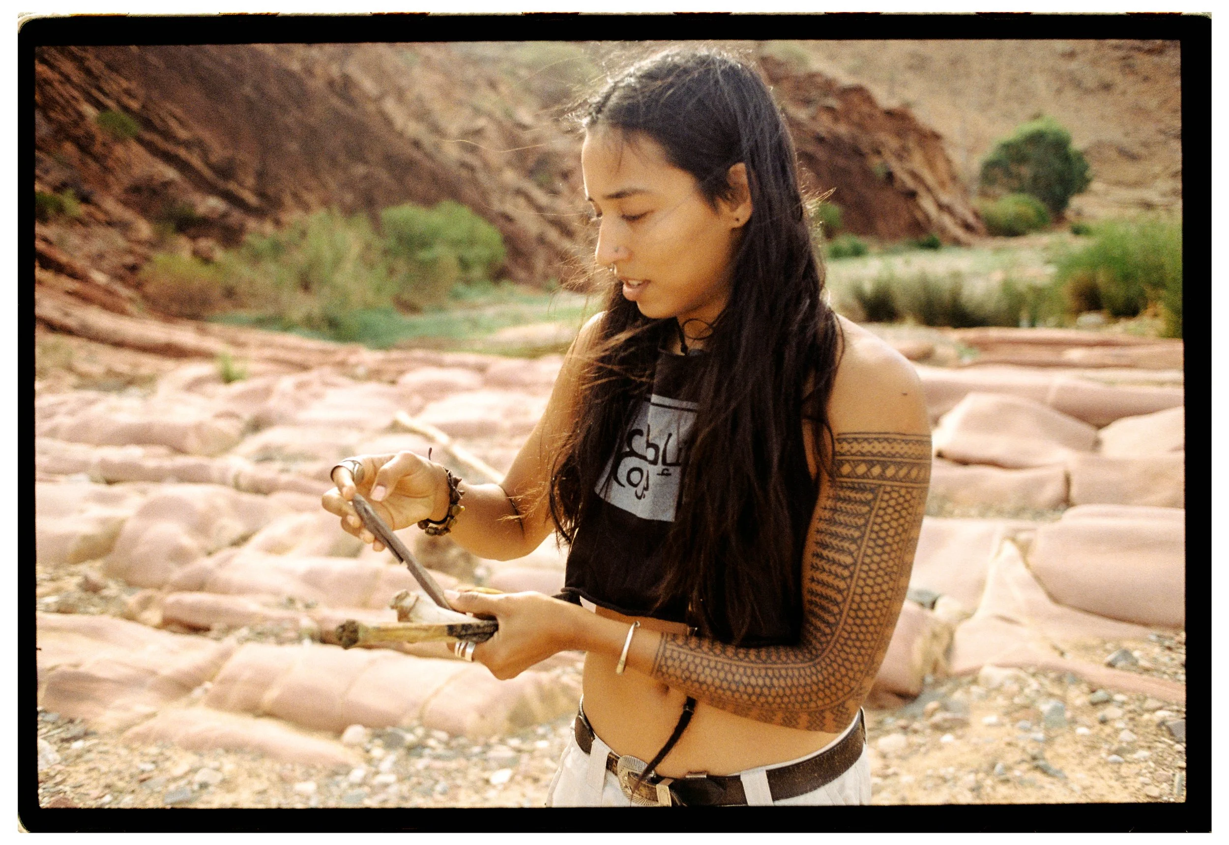 A woman with long dark hair, tattoos on her arm, wearing a bracelet and a ring, is looking at her phone while standing outdoors in a rocky desert landscape with red cliffs, small green bushes, and palm trees in the background.