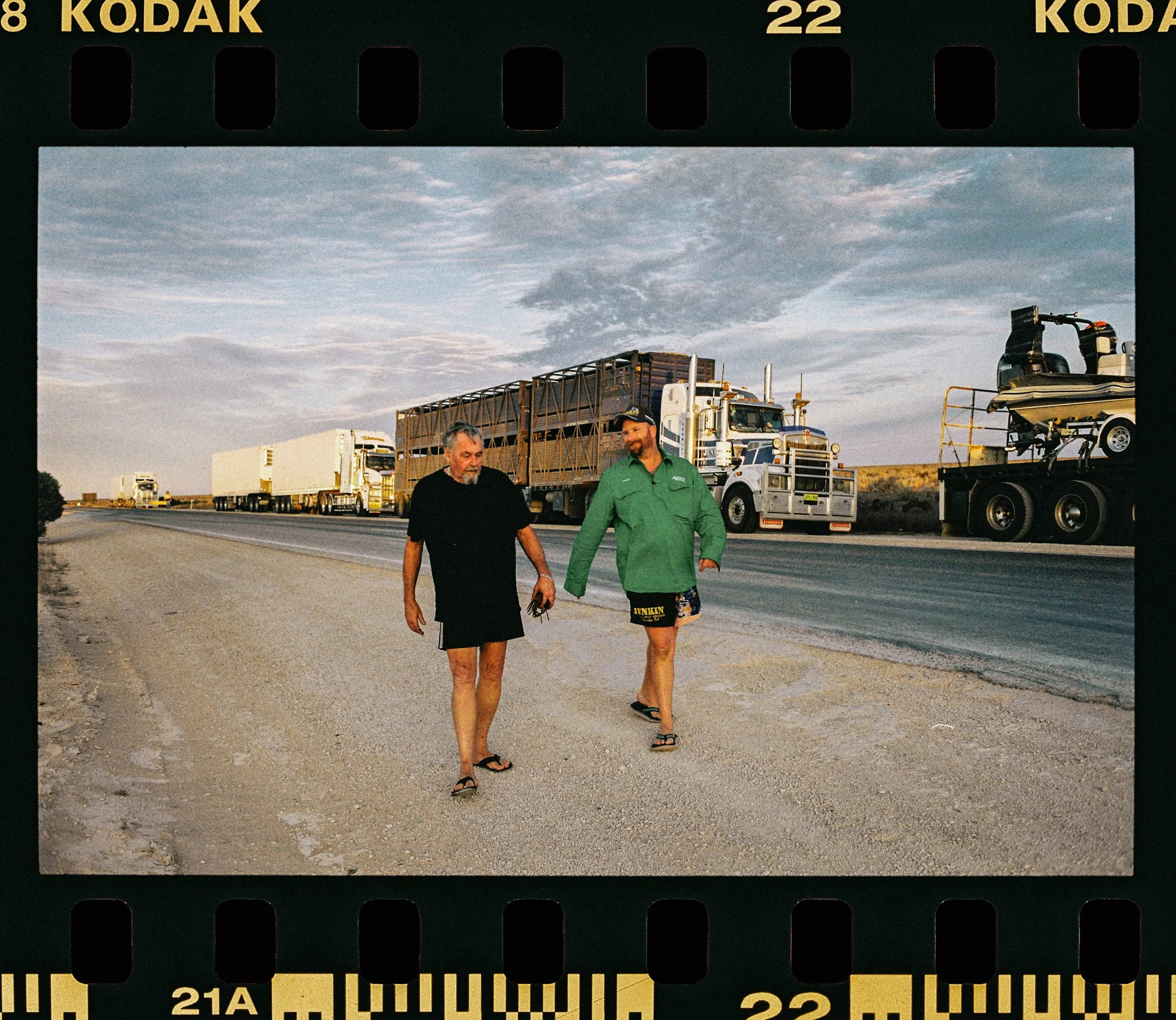 Two men walking on the side of a highway, with large trucks in the background and a vast sky overhead.