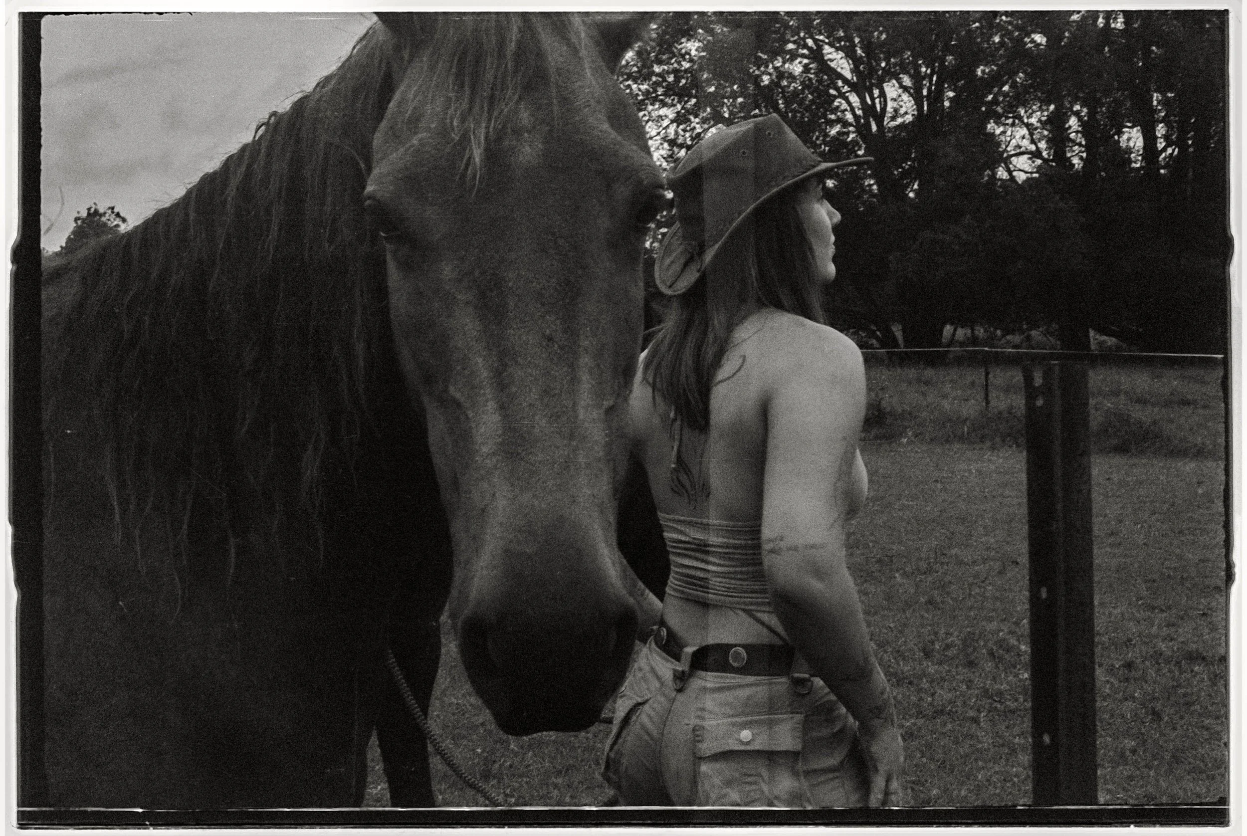 A woman wearing a cowboy hat standing next to a horse in an outdoor setting with trees in the background.