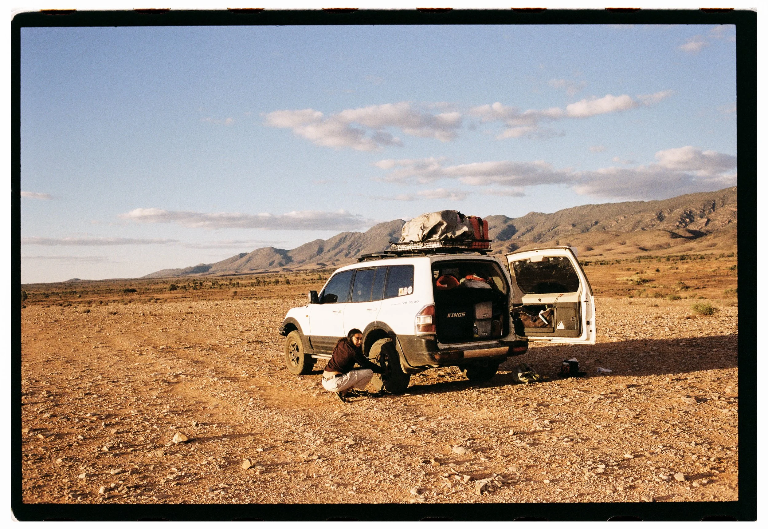 A man working on a car in a barren, rocky desert landscape with mountains in the background under a partly cloudy sky.