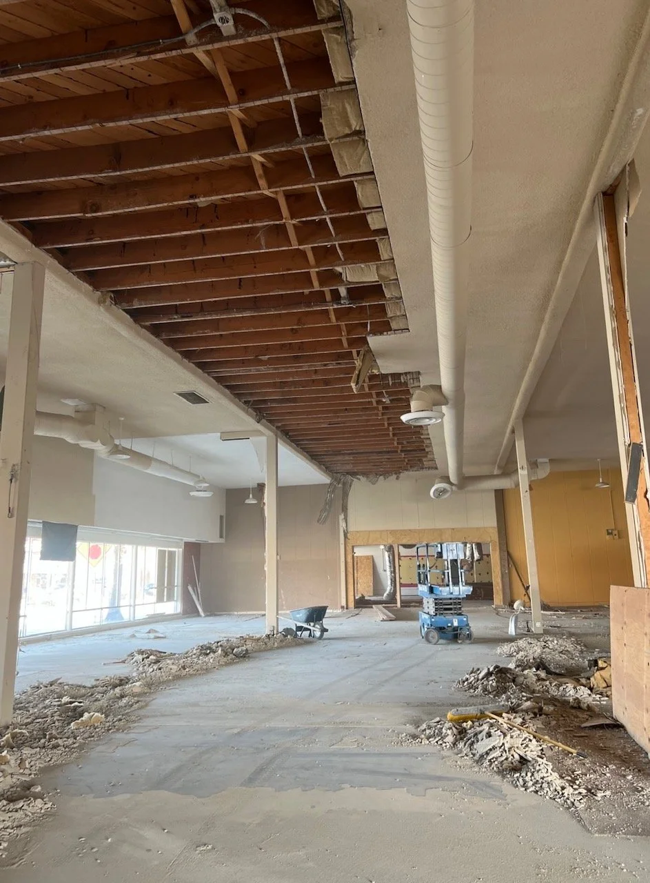 Interior of a building under renovation with exposed ceiling beams, visible ductwork, and construction debris on the floor.