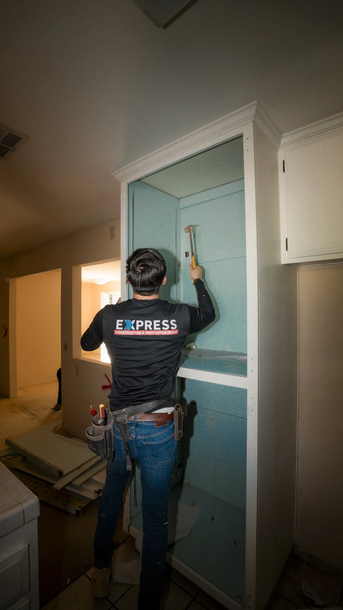A construction worker standing with his back facing the camera, using a hammer on a blue wall inside a cabinet or closet frame.