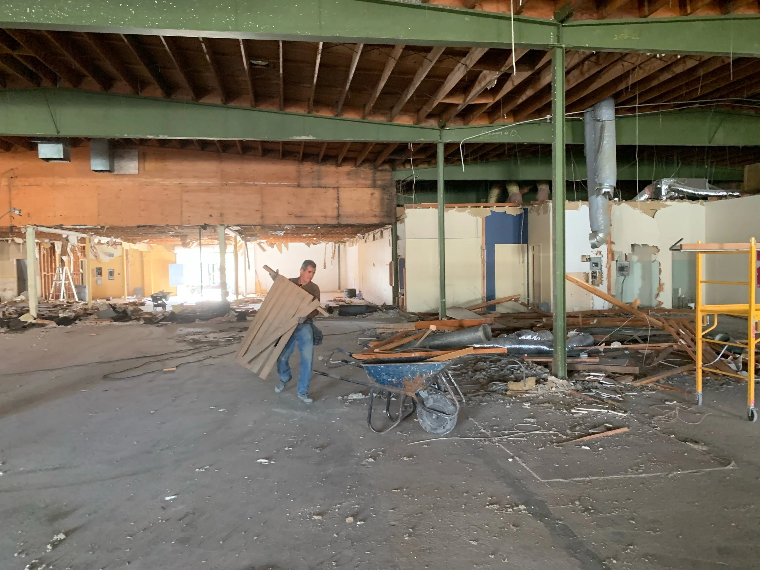 Interior of a building under renovation or demolition with a man holding wooden planks, construction debris scattered on the floor, a wheelbarrow, and construction tools and materials around.