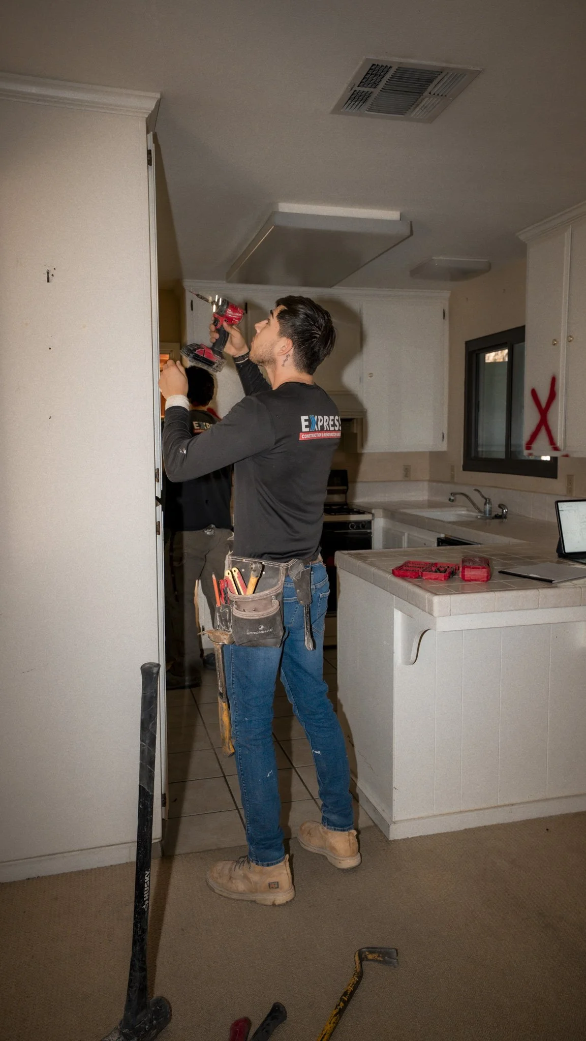 A handyman installing a ceiling vent in a kitchen, using a cordless drill, wearing work tools on his belt, with a few tools on the gray carpeted floor in front.