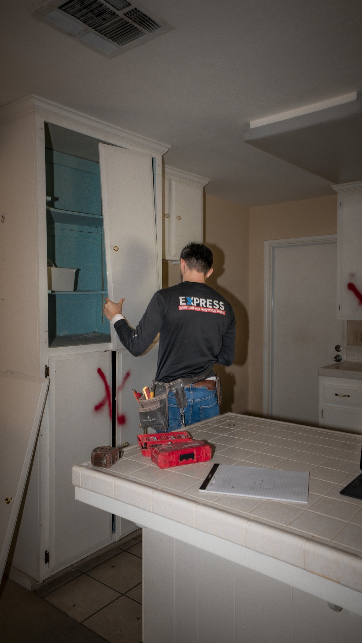 A construction worker with tools in a pouch on his belt is replacing a cabinet door in a kitchen with cream-colored cabinets, some of which are marked with red spray paint.
