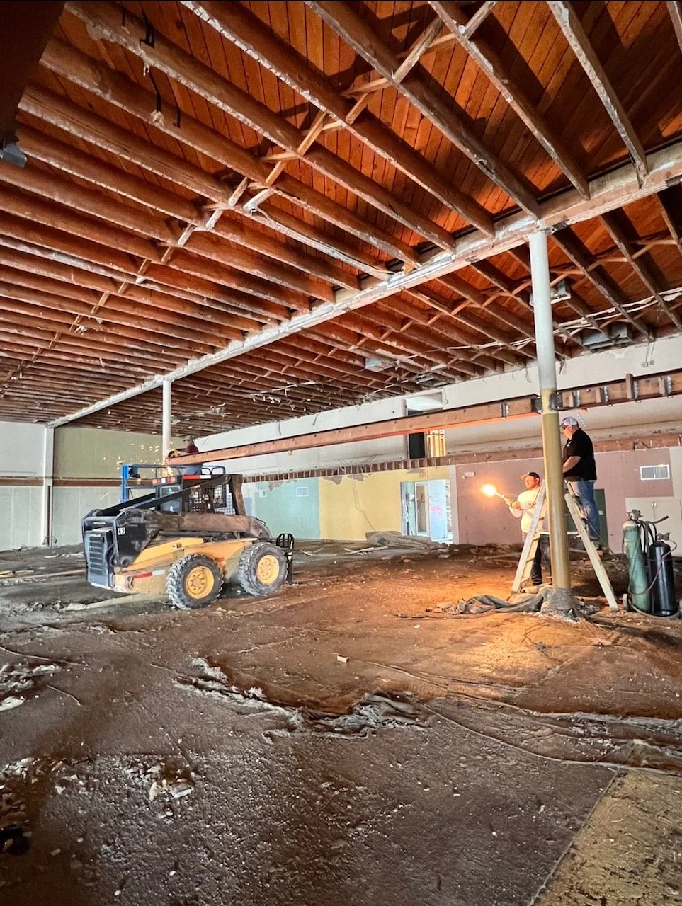Construction workers are working on a building's ceiling with exposed wooden rafters, using a lift and a ladder inside a partially renovated or demolished space with dirt and debris on the floor.