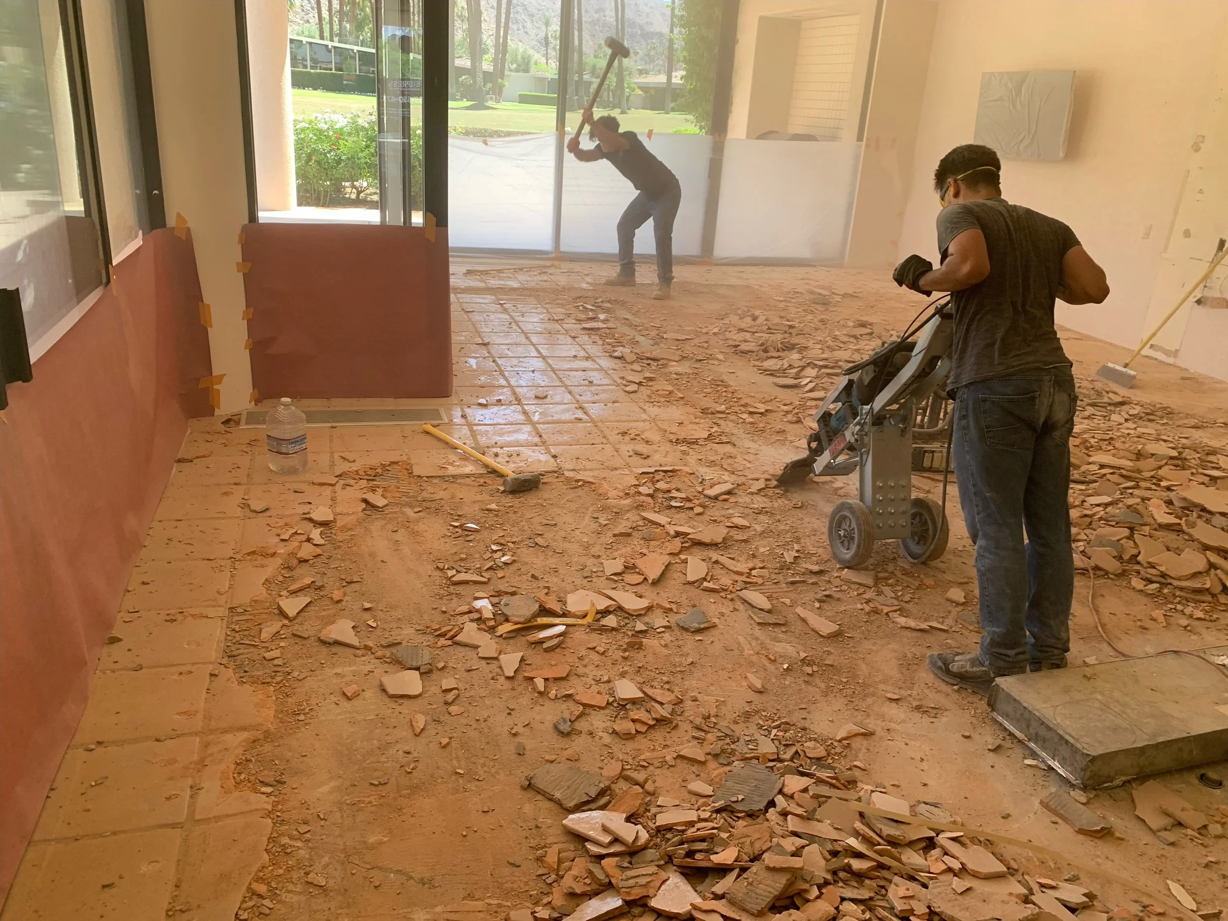 Two workers demolish a tiled floor inside a room with large windows and sliding glass doors, revealing an outdoor view with greenery.