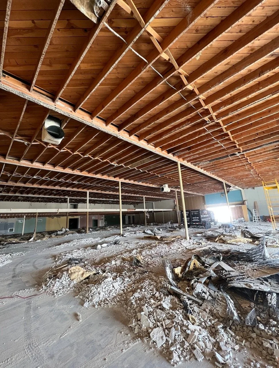 Construction or demolition site with exposed ceiling beams, debris, and construction equipment in a partially unfinished large indoor space.