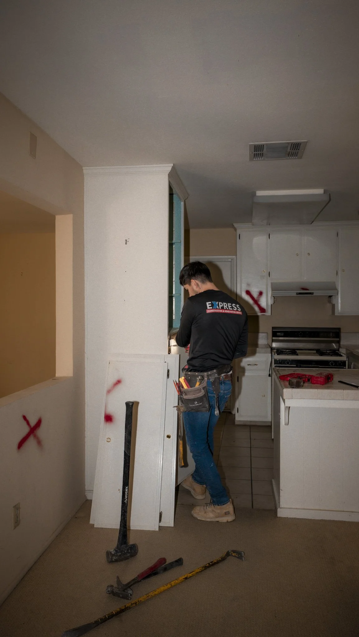 A construction worker with a tool belt stands in a kitchen, working on cabinet installation. The kitchen is partially renovated, with marked areas and tools on the floor.