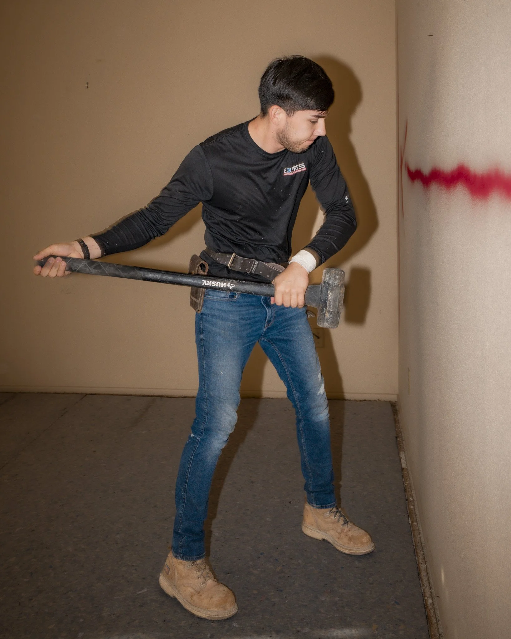 Person wearing black long-sleeve shirt, blue jeans, and tan work boots, hitting a wall with a sledgehammer.