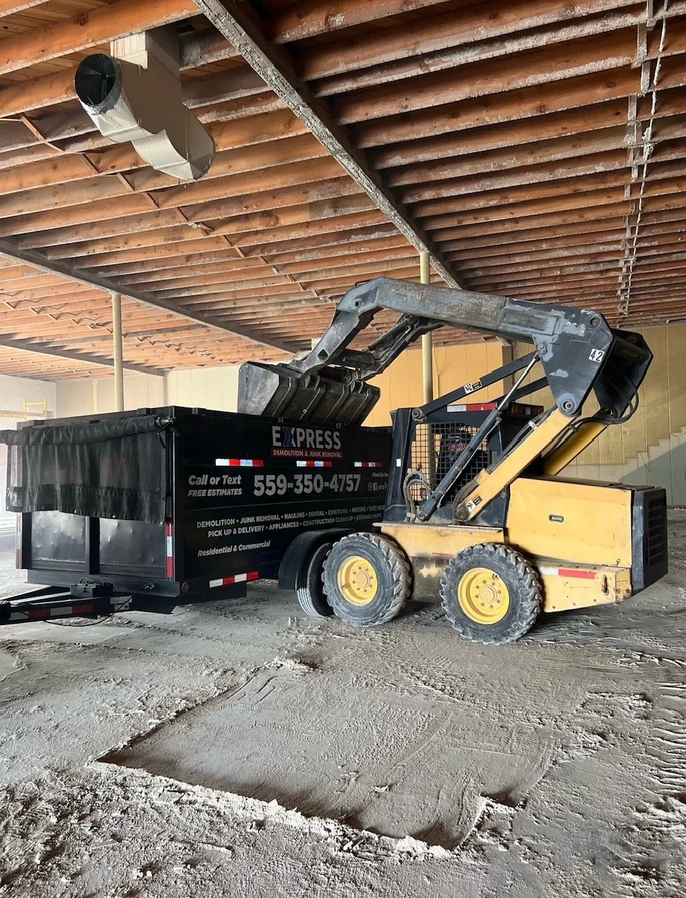 Construction site with a small skid-steer loader dumping dirt into a black trash bin. The area has unfinished ceilings with exposed wooden beams and ductwork.