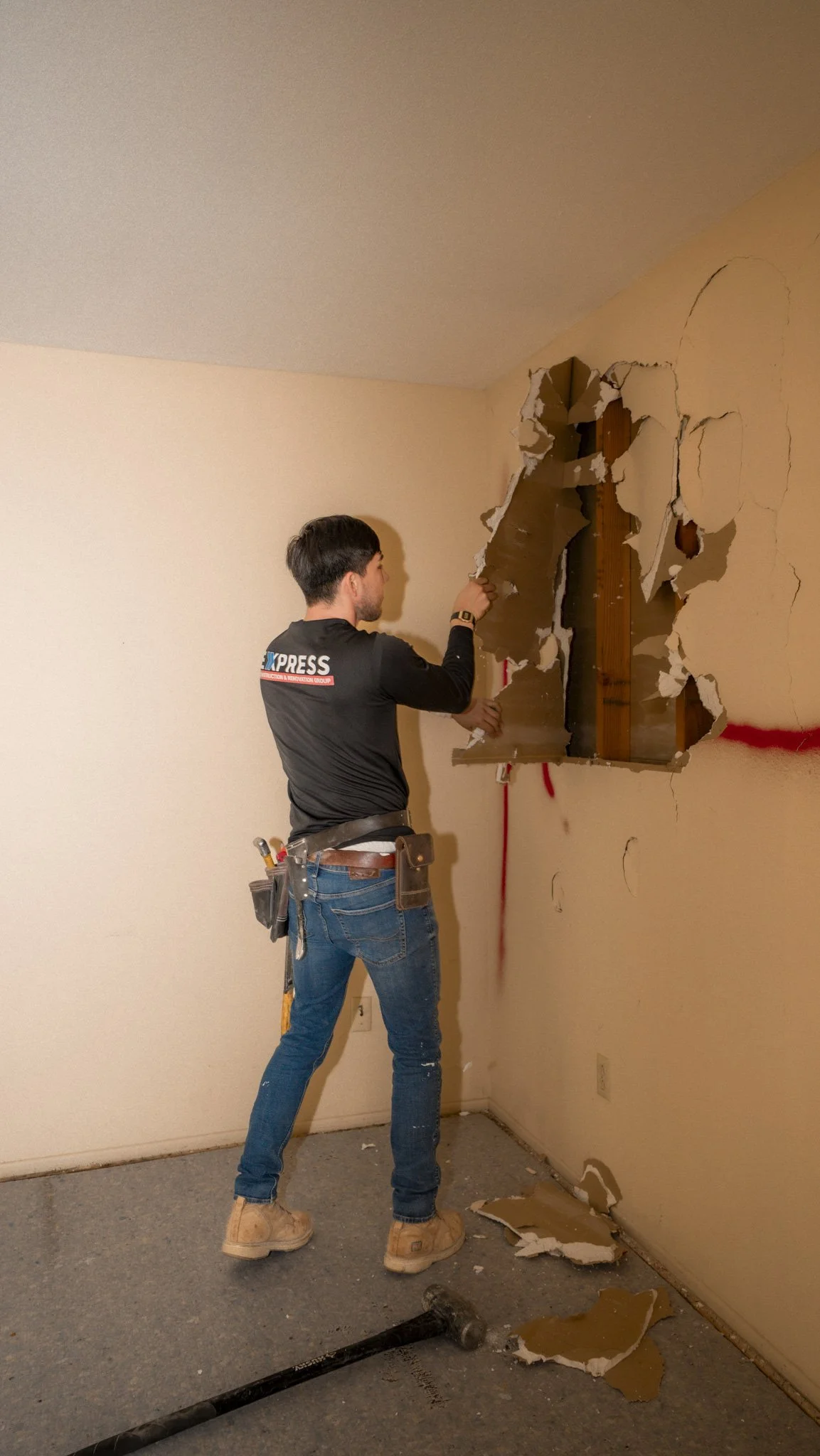 A worker in black clothes and blue jeans breaking through a drywall wall with a hammer, with debris on the carpeted floor.