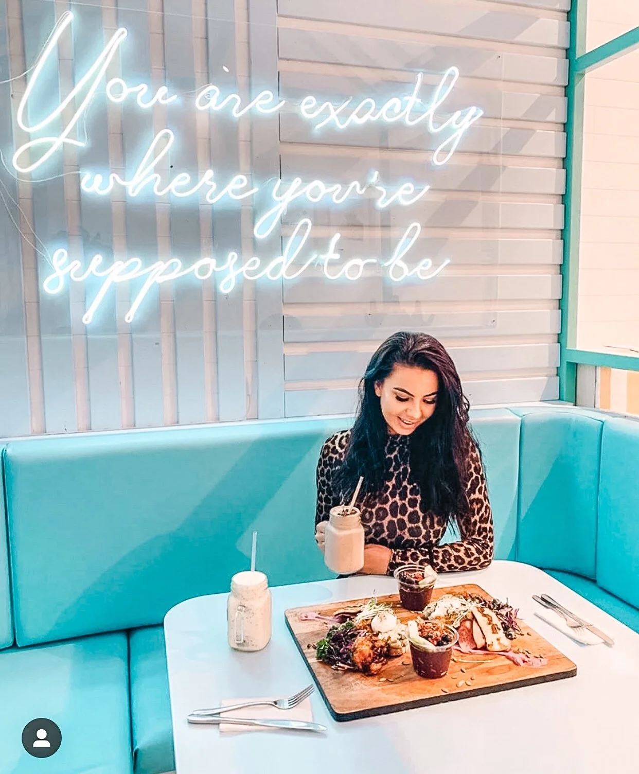 A woman with dark, curly hair in a leopard print top sitting at a white table with a turquoise booth seating. She is holding a Mason jar drink, surrounded by food on a wooden board, with cutlery and condiments on the table. Behind her, a neon sign sa