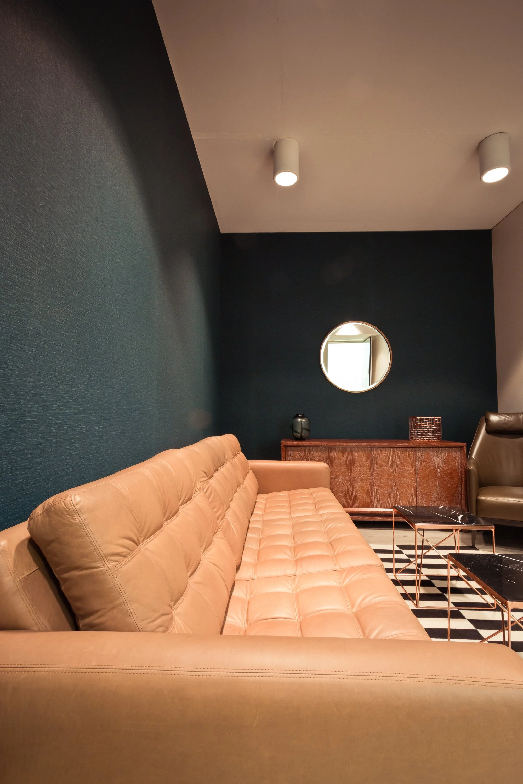 Modern living room with tan leather tufted sofa, black wall, round mirror, wooden sideboard, and black marble coffee tables.