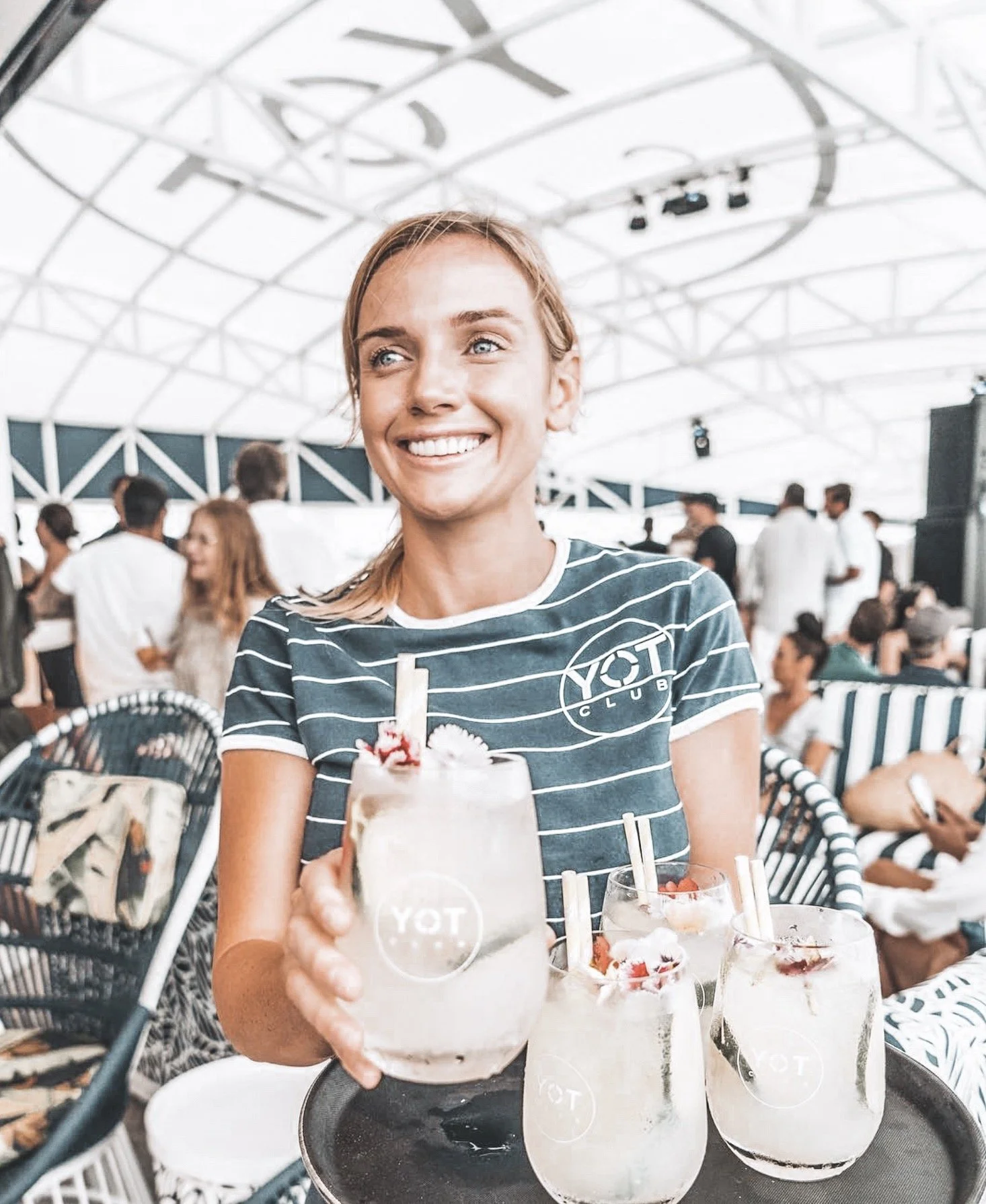 A woman smiling and holding a tray with three large cocktails at a social gathering.
